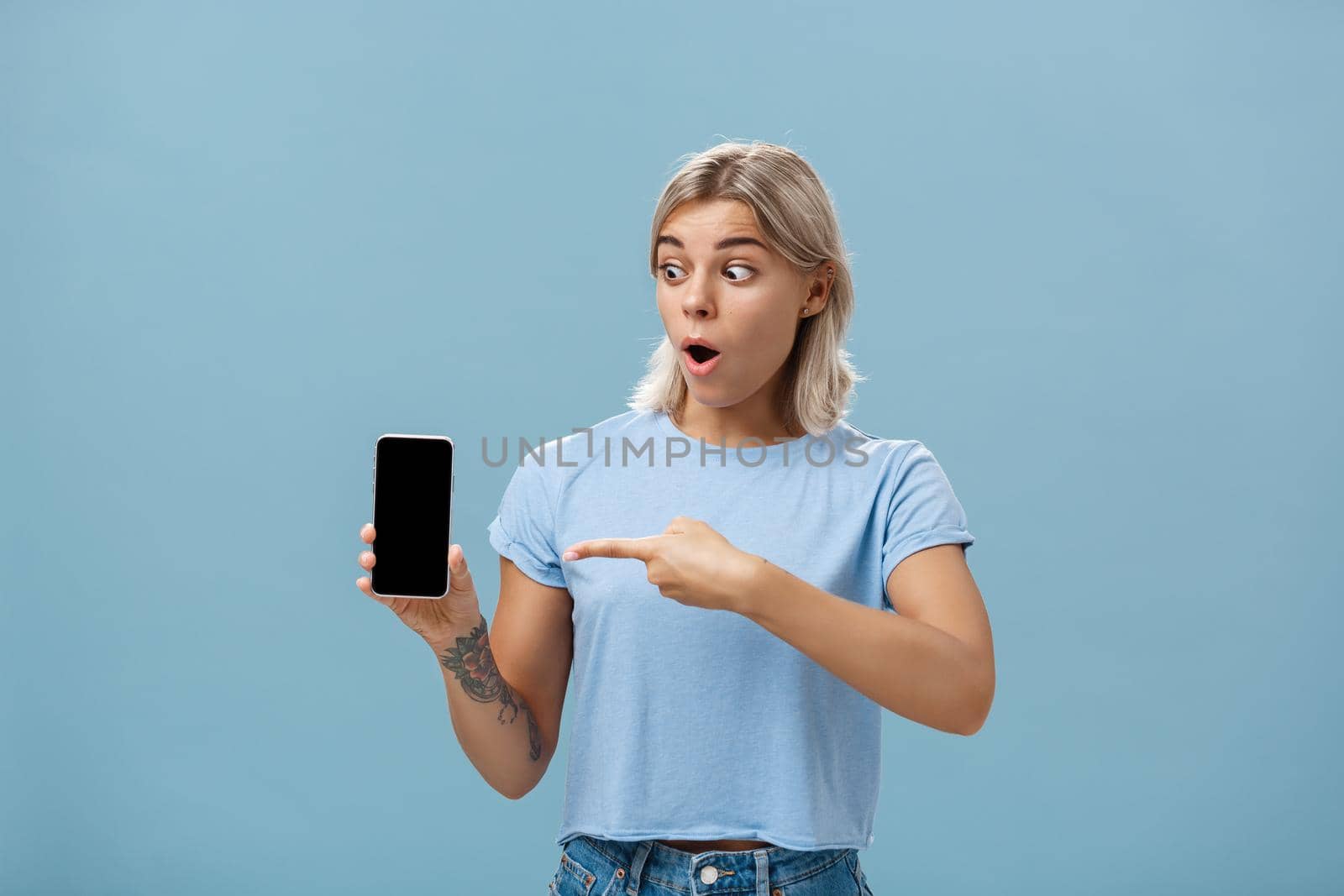 Girl being amazed with cool new smartphone received on birthday. Surprised speechless happy woman with fair hair and tattoo dropping jaw holding smartphone pointing and staring at gadget screen.