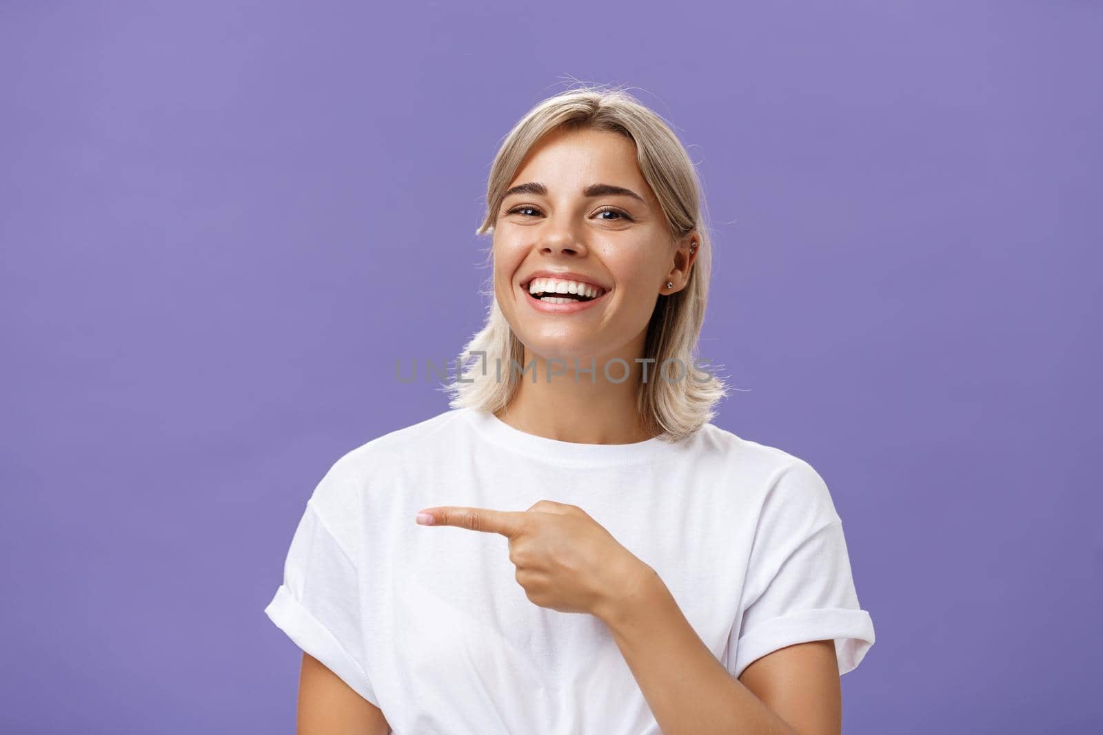 Close-up shot of amused happy and entertained good-looking sociable woman with fair hair and beautiful smile grinning while pointing left with index finger showing awesome copy space over purple wall by Benzoix