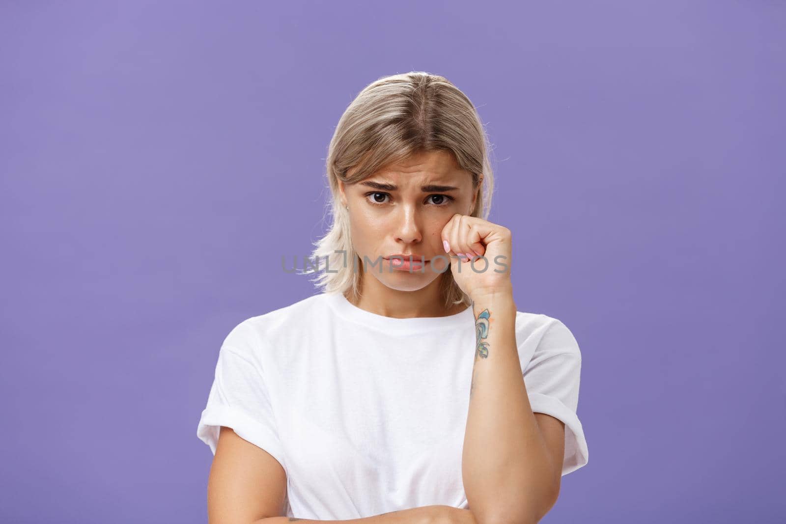 Studio shot of offended sad and timid silly woman with blond hairstyle frowning looking from under forehead holding fist near eye as if whiping teardrop being upset over purple background by Benzoix