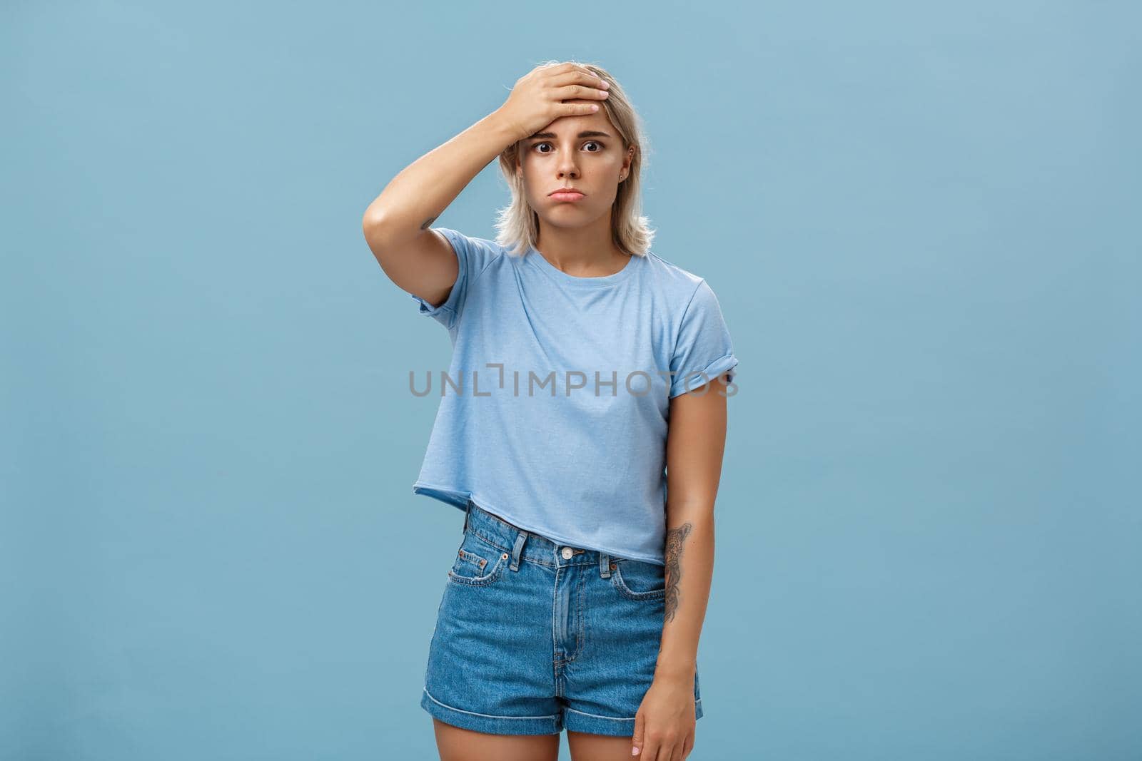 Concerned and troubled young teenage girl being shocked with loads of responsibilities holding hand on forehead staring spaced out and pouting from tiresome perplexed situation over blue wall by Benzoix