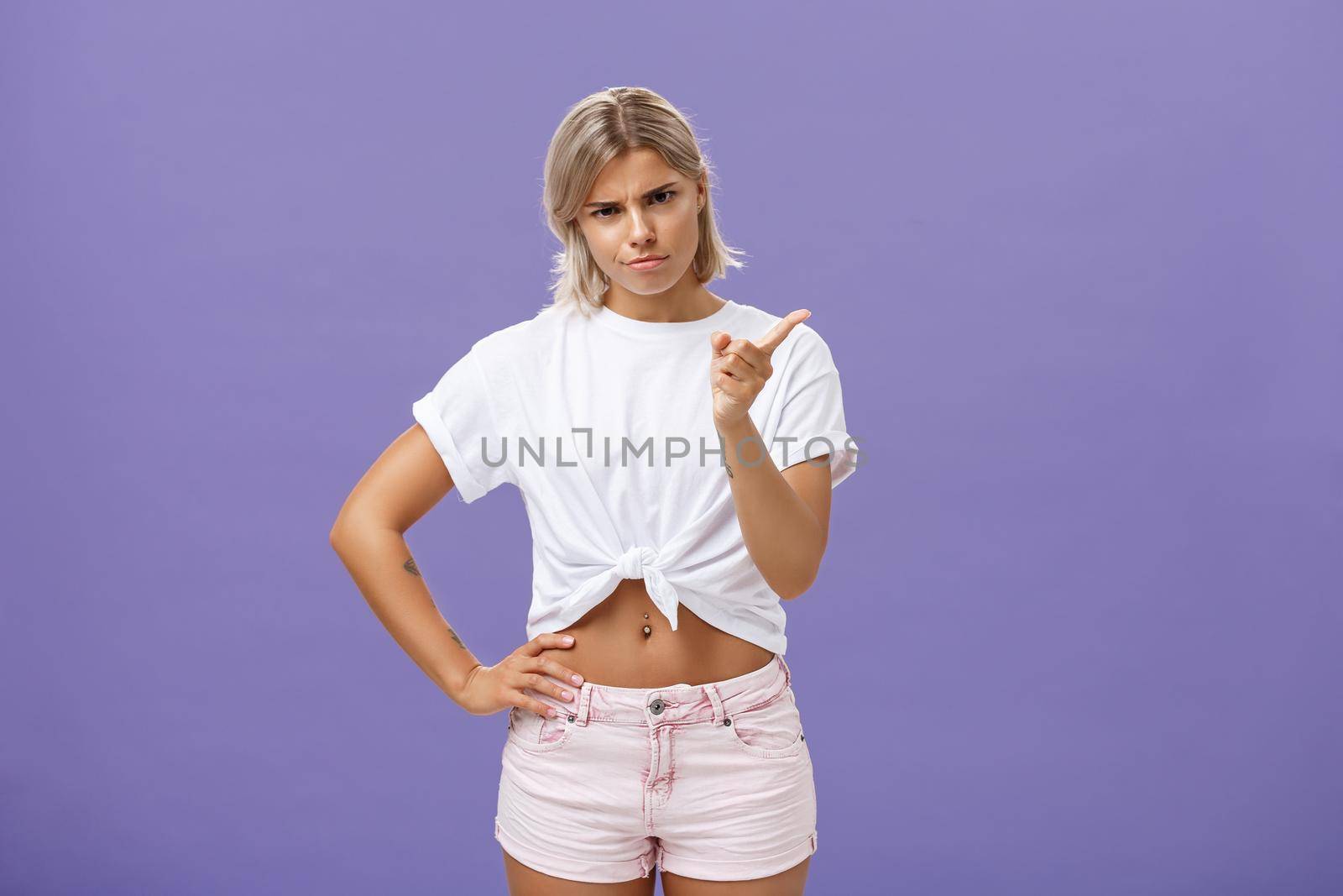 I told you behave properly. Strict and cute displeased blonde female in white t-shirt and shorts shaking index finger in strict pose looking from under forehead while scolding over blue background by Benzoix