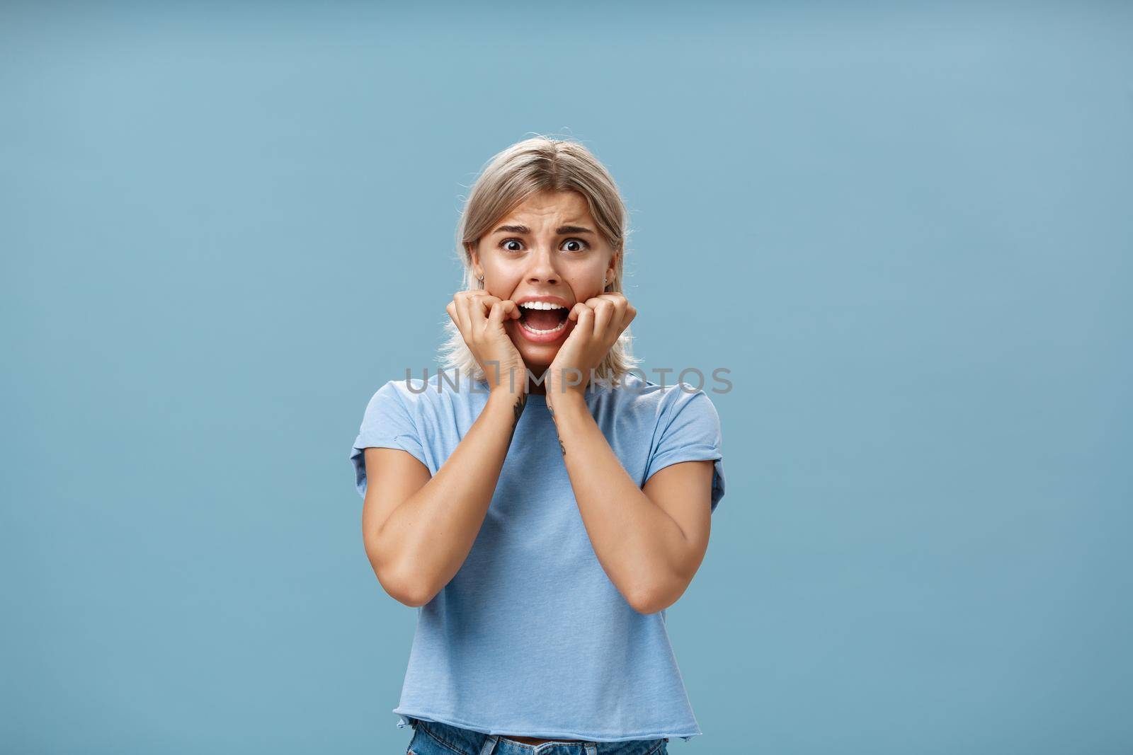 Indoor shot of shocked troubled girlfriend hearing mom returned home being with boyfriend biting fingernails being in helpless perplexed situation feeling anxious and scared over blue background by Benzoix