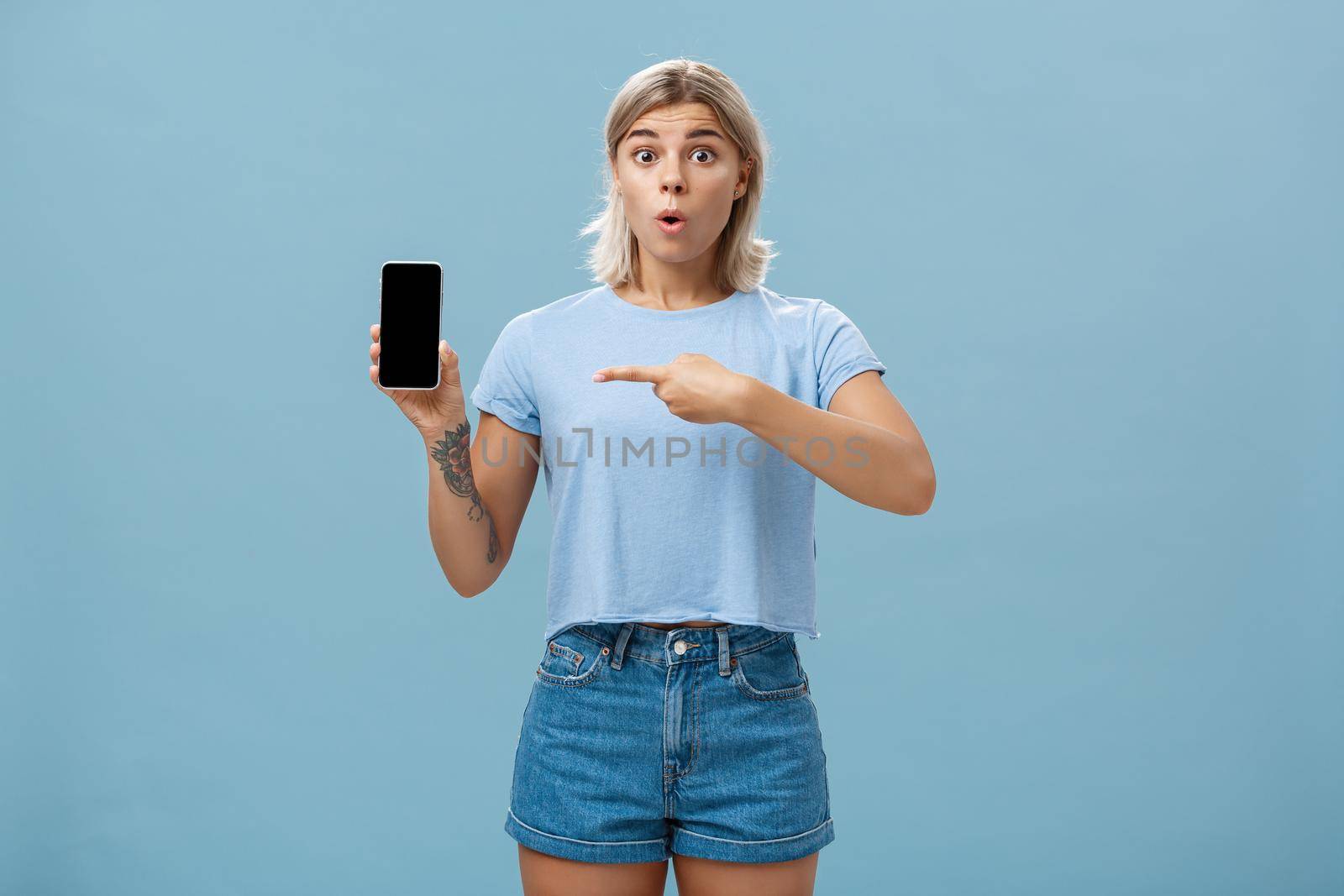 Wow look what found online. Portrait of impressed speechless good-looking young sportswoman in t-shirt and denim shorts saying wow folding lips showing smartphone and pointing at screen over blue wall by Benzoix