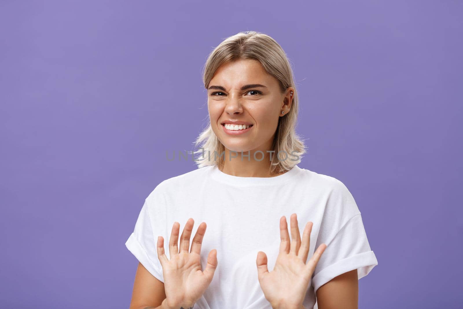 Nah no thanks. Unimpressed cute stylish woman unwilling accept offer raising palms in no rejection gesture clenching teeth while smiling with intense displeased smile frowning over purple background by Benzoix