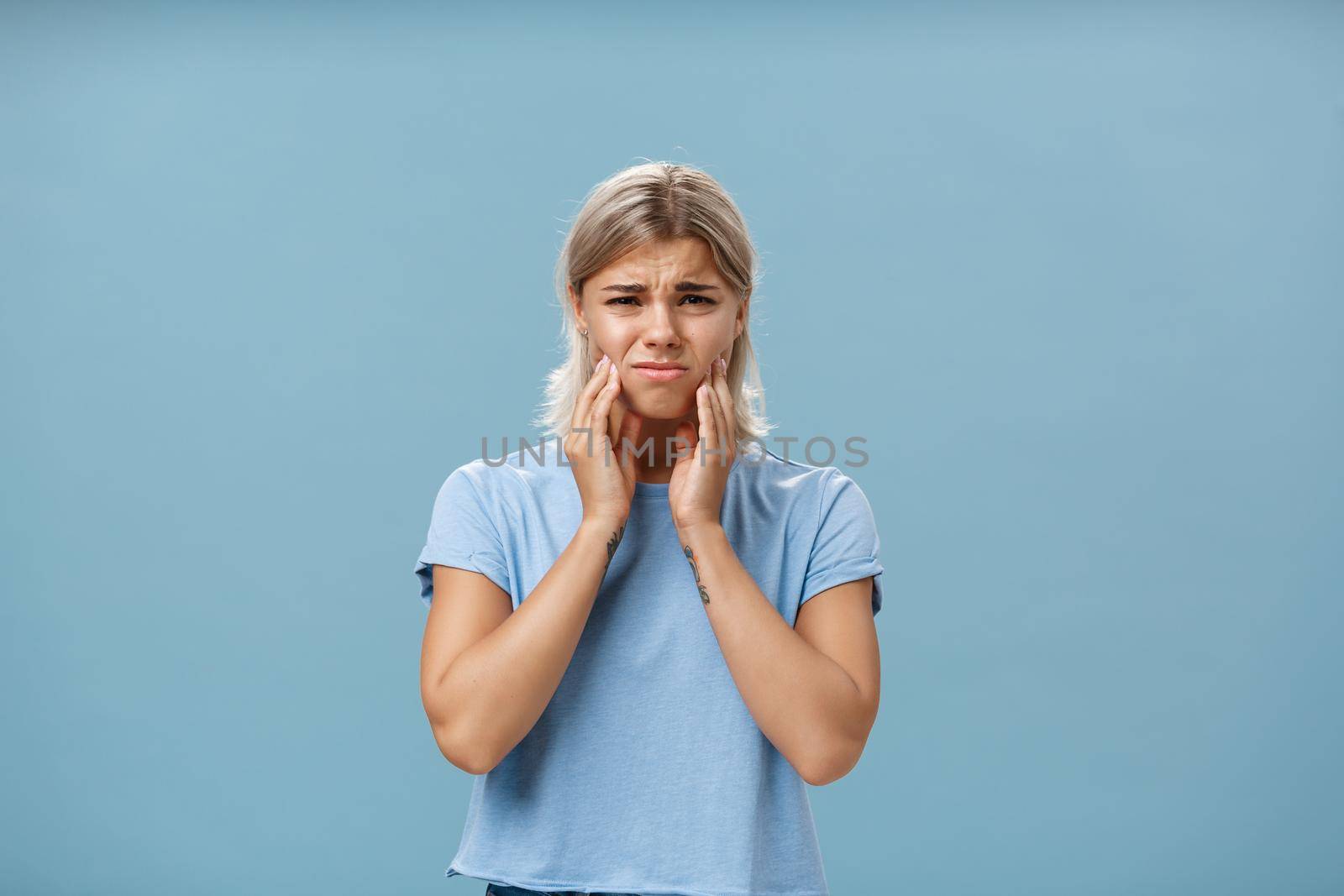 Girl feeling pain in teeth after drinking hot coffee. Displeased unhappy troubled young girl with blond hair frowning feeling discomfort in mouth holding hands on cheeks suffering toothache by Benzoix