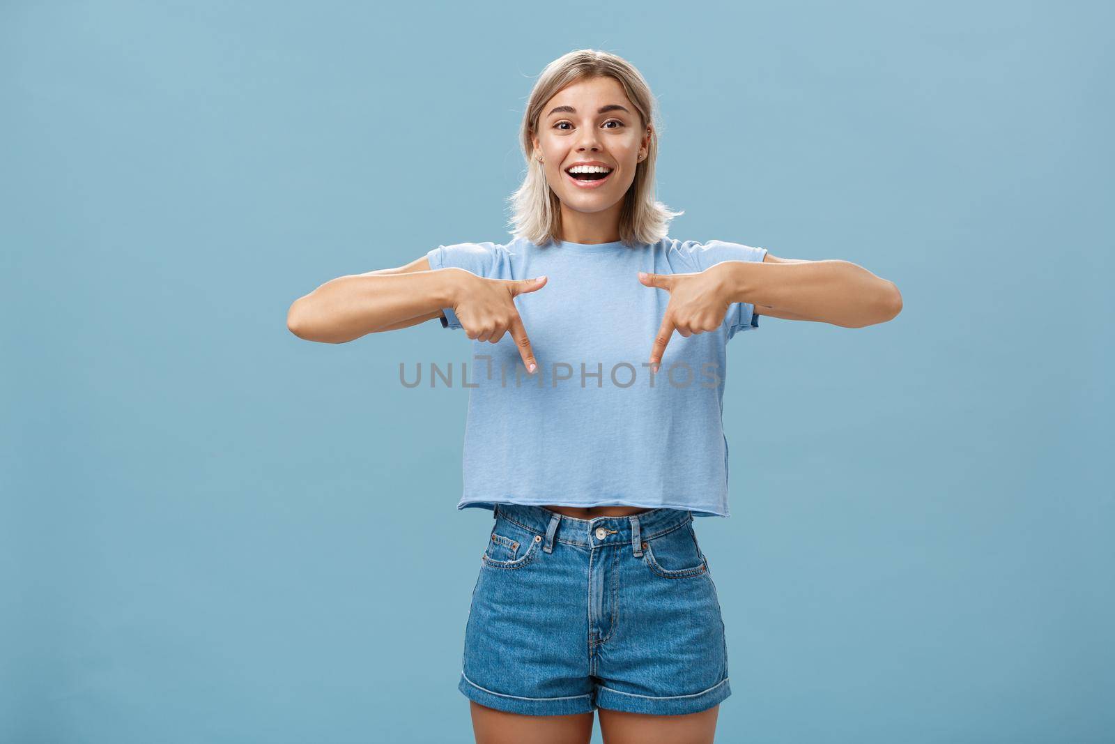 You have to see this. Portrait of joyful amused and happy stylish blonde female in trendy t-shirt pointing down with arms near chest smiling broadly showing amazing copy space over blue background by Benzoix