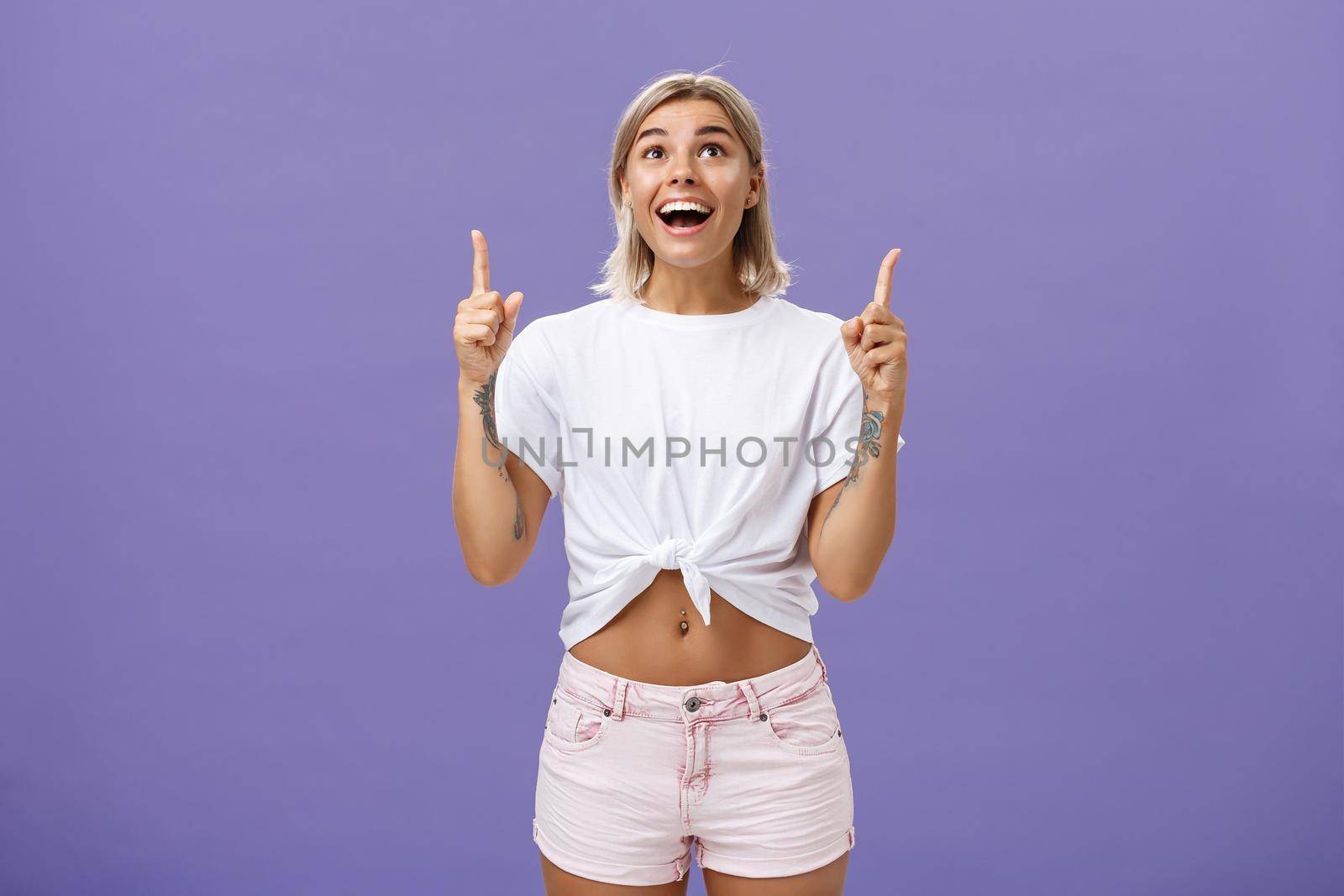 Woman seeing miracle being impressed and delighted like child staring and pointing up with broad thrilled smile standing over purple background in summer trendy outfit being joyful and excited by Benzoix