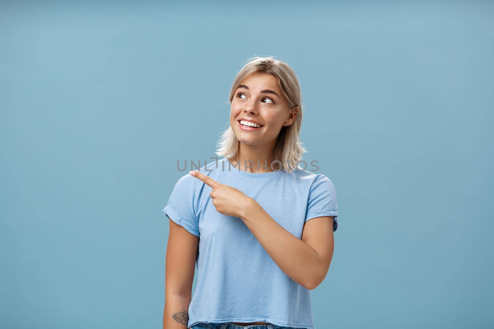 Waist-up shot of creative and dreamy happy attractive blonde female in casual t-shirt looking and pointing at upper left corner with admiration and joy standing over blue background amused and curious by Benzoix