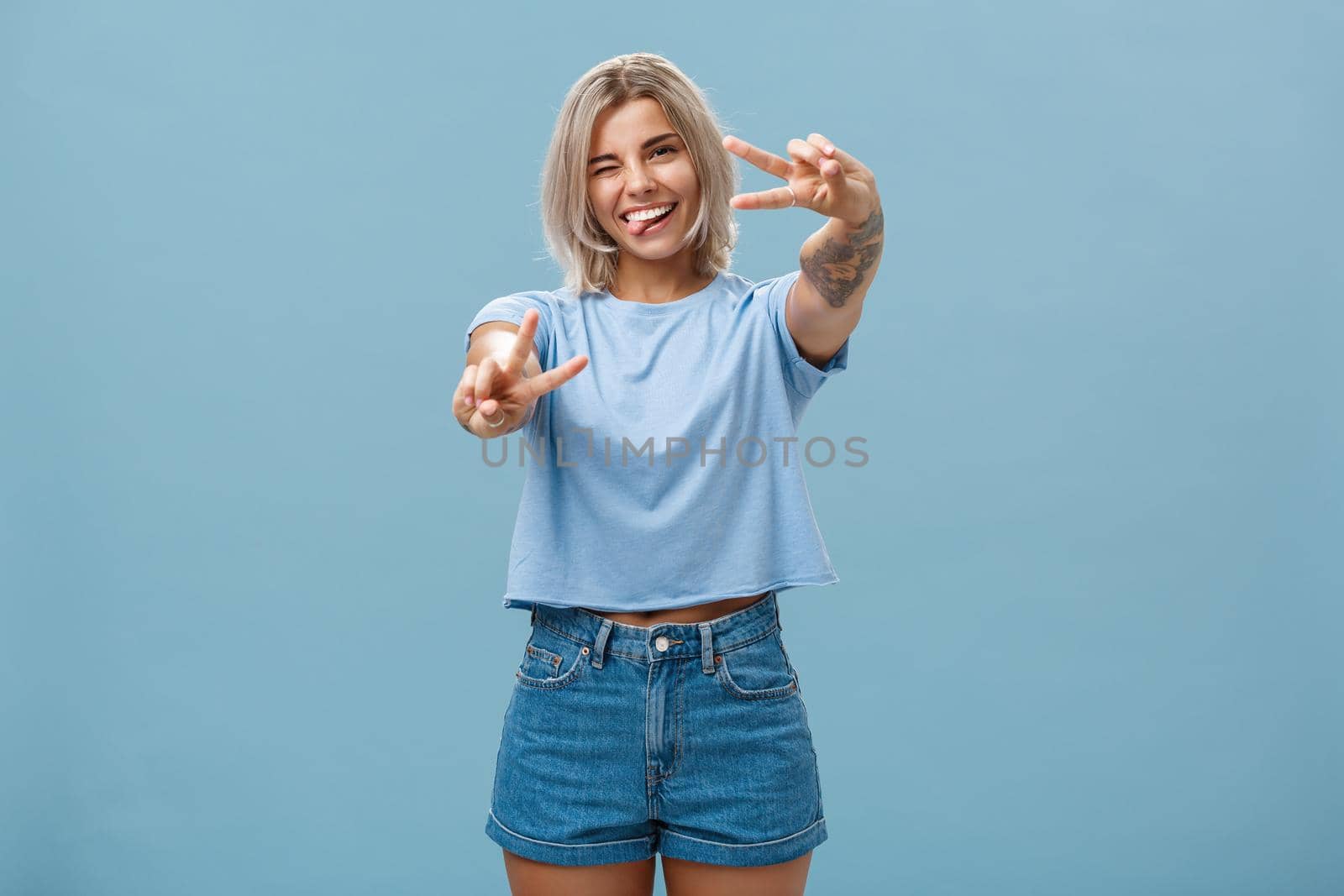 Beautiful tattooed girl enjoying weekends sticking out tongue joyfully winking at camera and smiling showing peace or victory gesture with pulled arms feeling happy posing over blue background by Benzoix