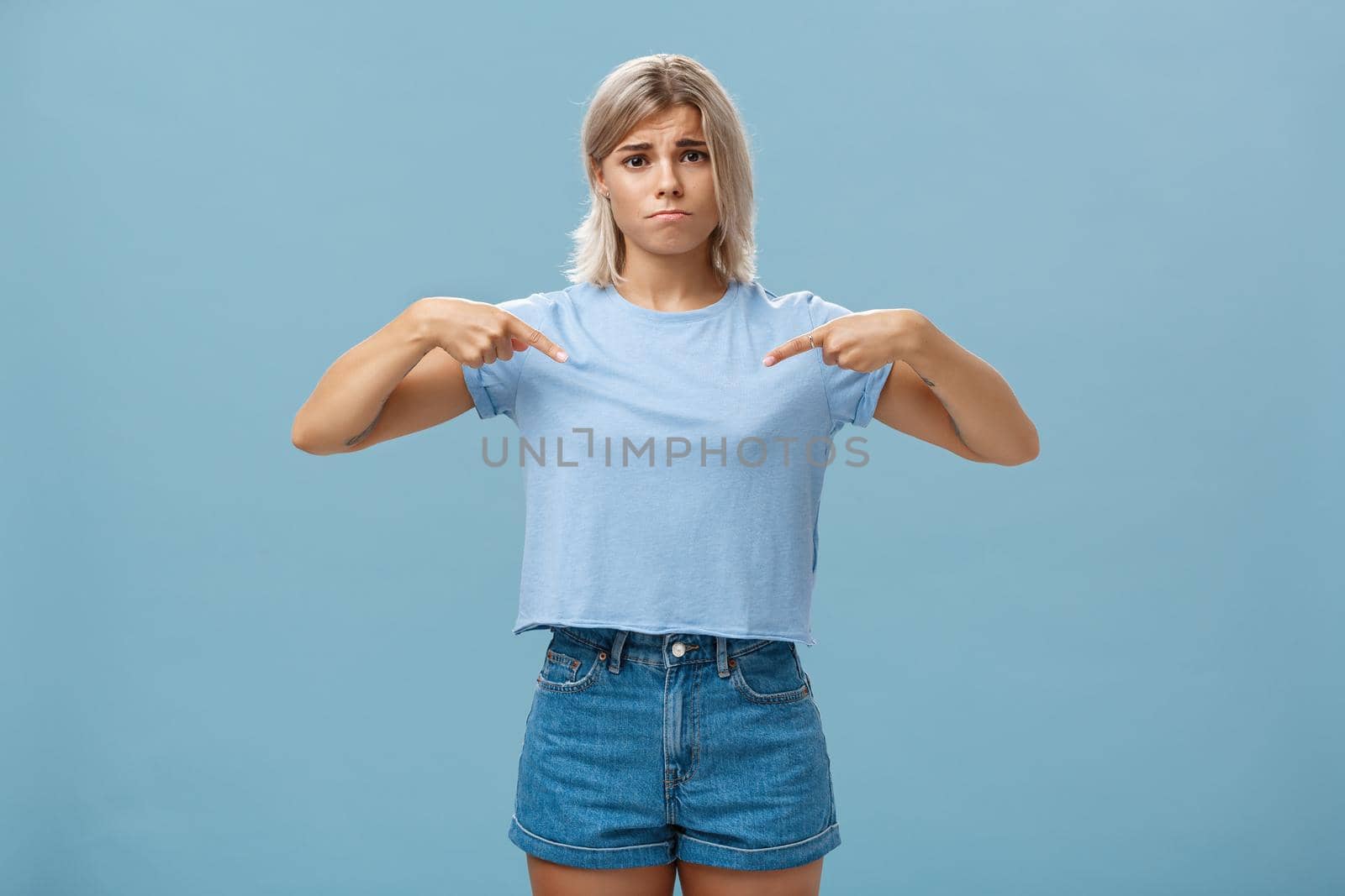 Sad cute blonde young female complaining being displeased with own body, pointing at breast frowning making gloomy smile pulling lips down from disappointment standing over blue background.