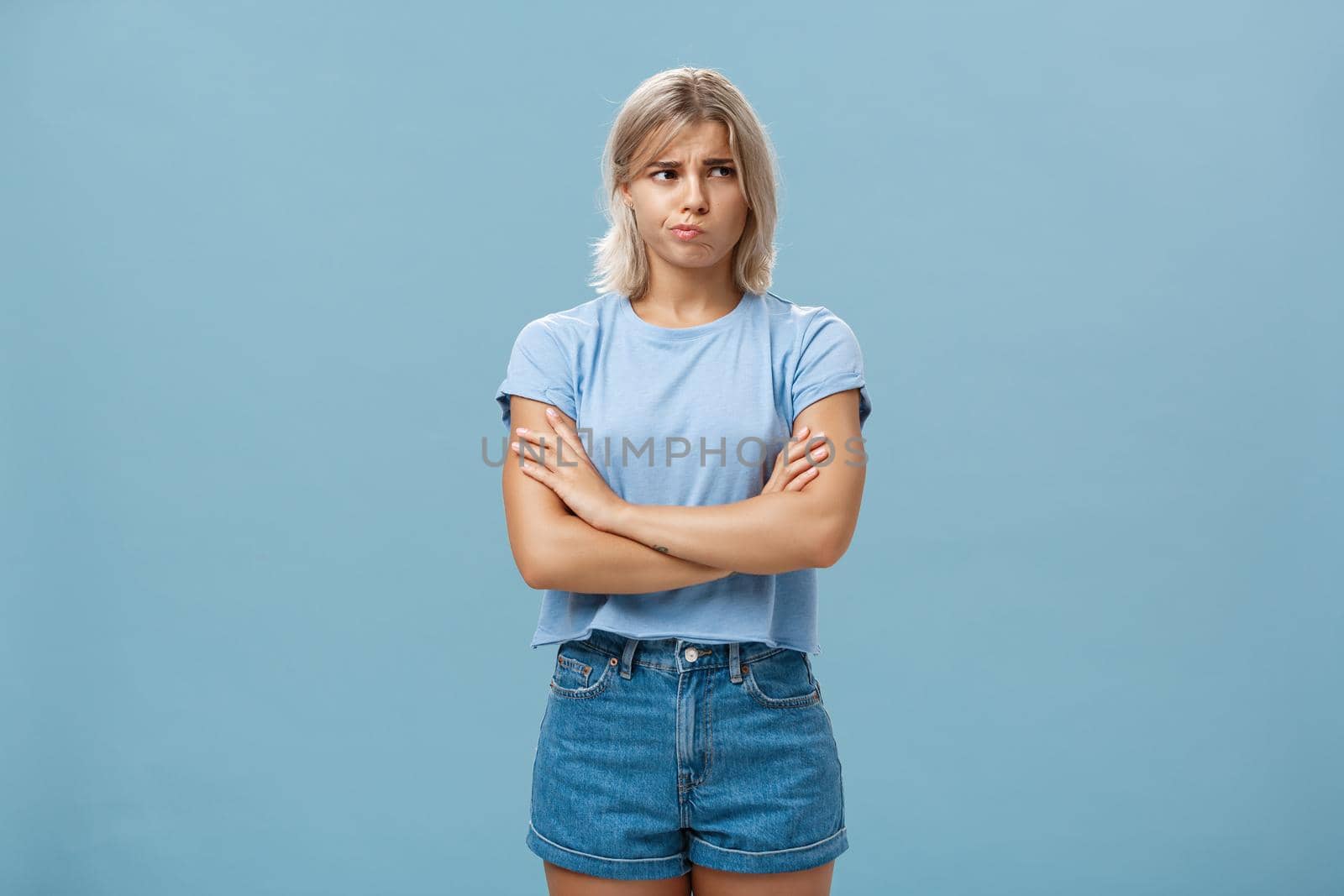Uncertain troubled and perplexed attractive blond woman with tanned skin holding hands crossed on chest pouting and frowning looking right with worried unsure and doubtful look over blue wall by Benzoix