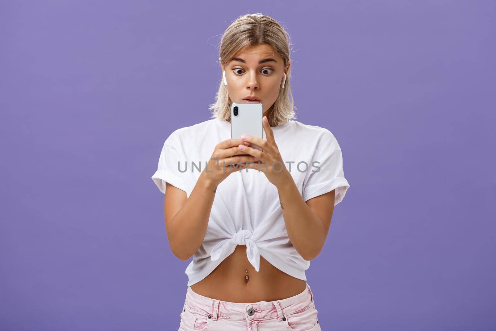 Girl watching thrilling video on social media in wireless earphones holding smartphone near face popping eyes at smartphone screen gazing at gadget with interest and curiosity over purple background. Technology concept