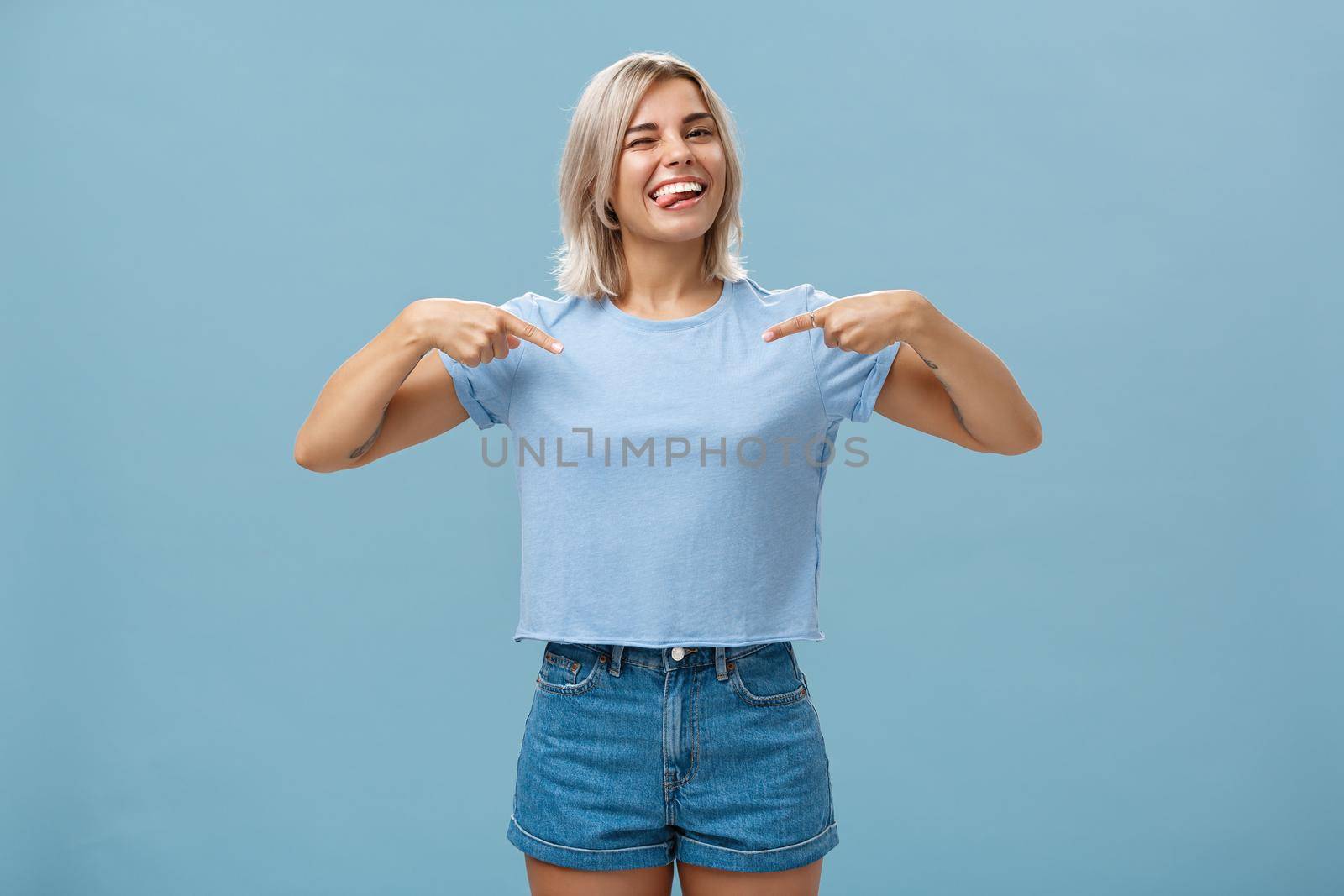 Yeah proud of myself. Self-assured joyful and carefree attractive blonde female with tanned skin and tattoos pointing at herself winking and sticking out tongue with broad smile over blue wall by Benzoix