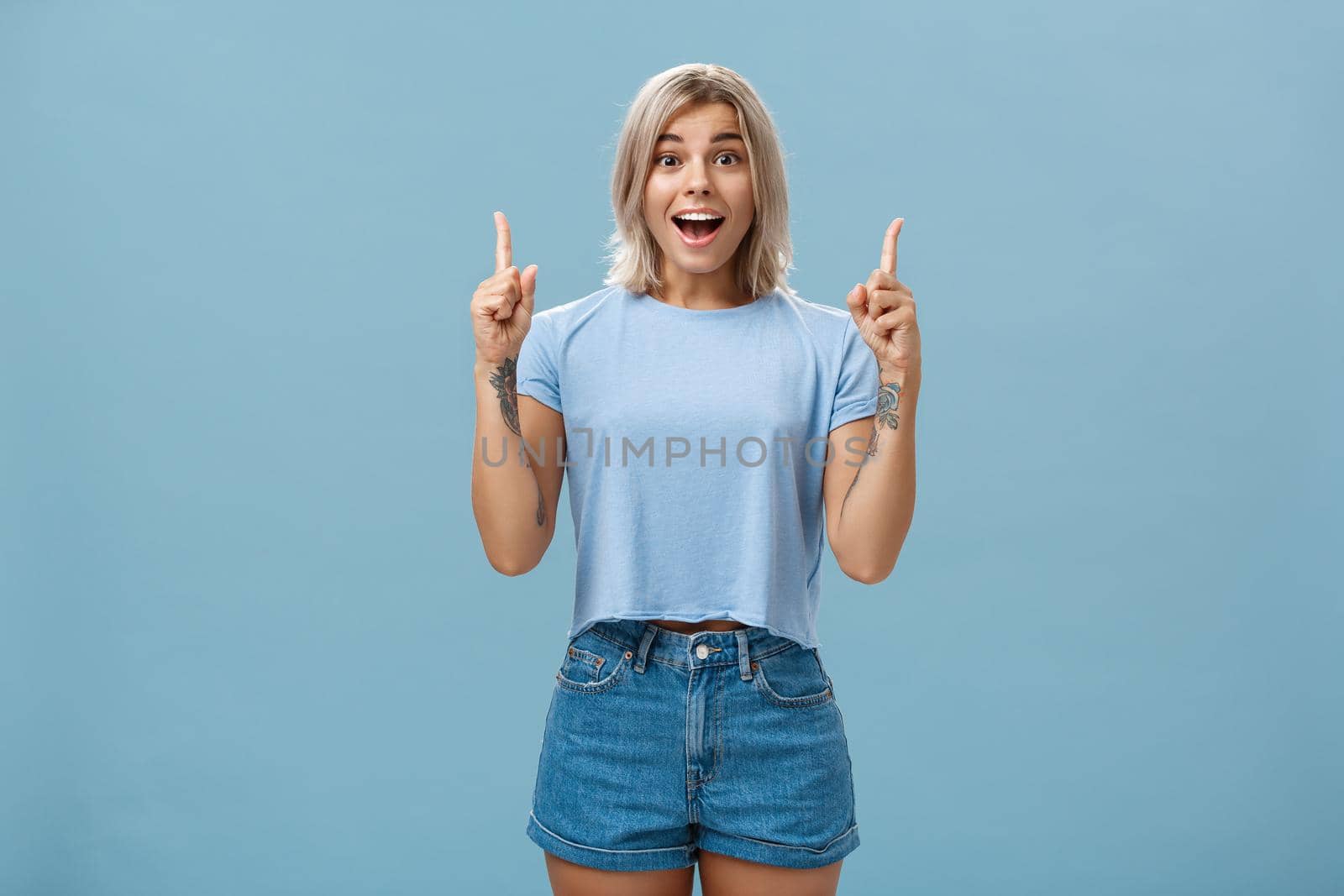 Waist-up shot of impressed enthusiastic creative blonde woman in trendy summer outfit smiling gasping being charmed and thrilled with awesome copy space pointing up over blue background. Copy space