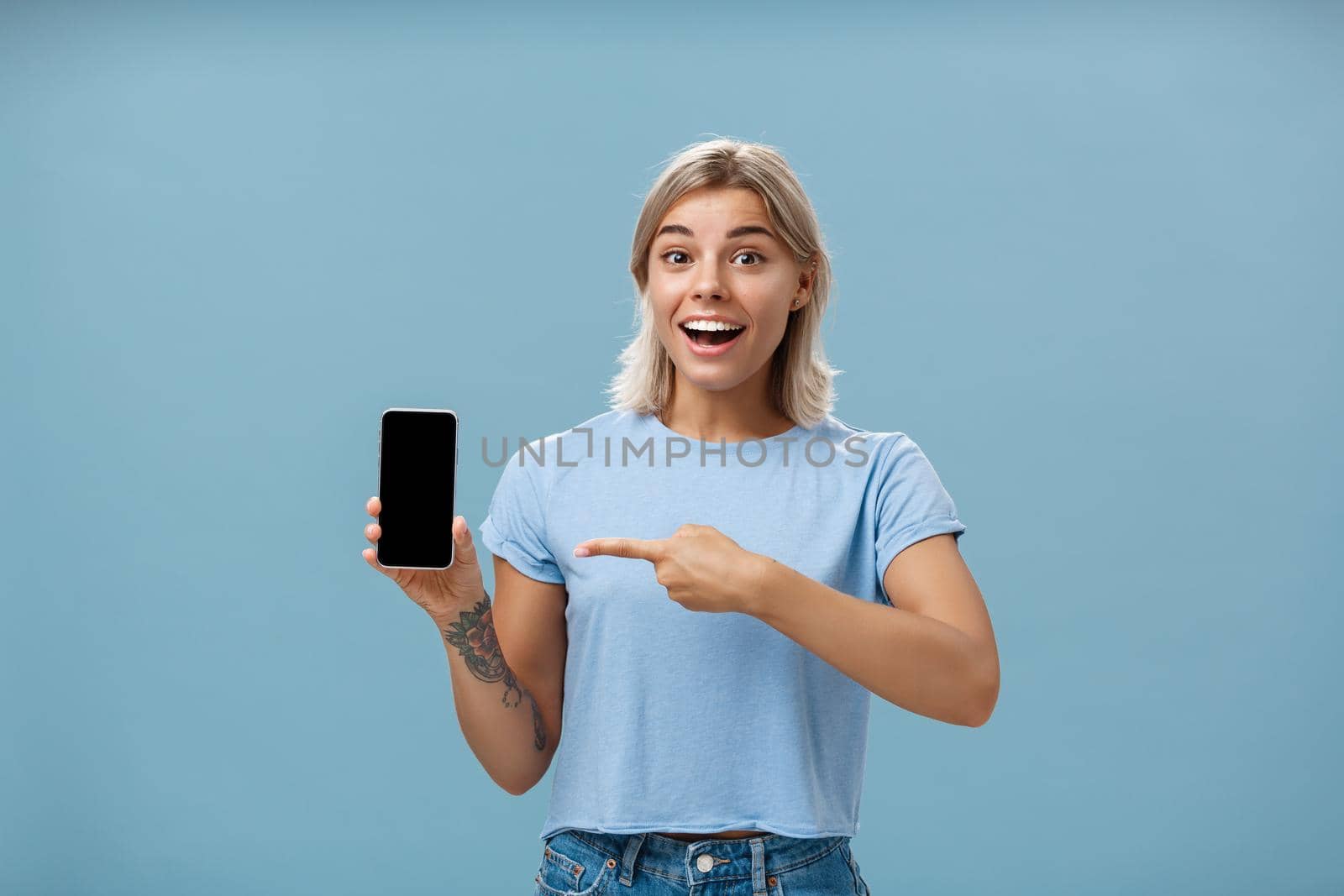 Waist-up shot of thrilled and impressed good-looking female student in casual t-shirt smiling joyfully pointing at smartphone screen showing awesome place via internet to friend over blue background by Benzoix