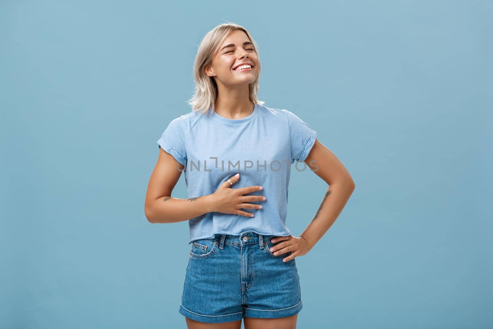 Cute girl likes eat after workout being pleased and stuffed after dinner. Portrait of satisfied attractive blonde young woman touching belly raising head up with broad smile over blue background.