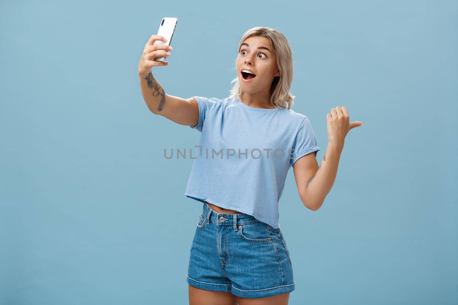 Lifestyle. Girl recording video showing followers awesome scene attending cool event holding smartphone looking impressed and surprised at device screen pointing backwards indicating right over blue wall.