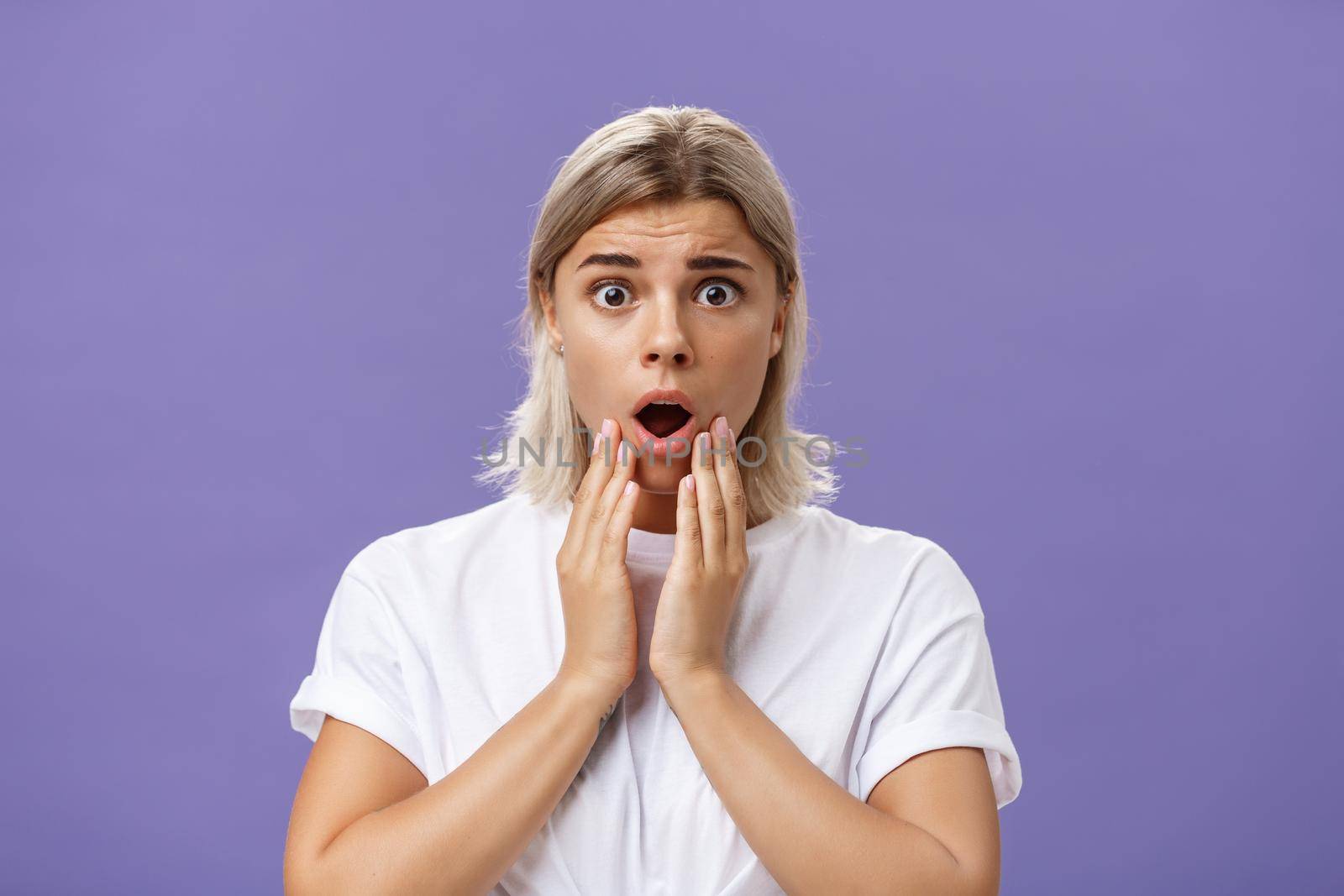 Waist-up shot of worried and shocked woman expressing empathy frowning holding hands near opened mouth feeling sorry and nervous hearing terrible disaster happened standing over purple wall. Emotions concept