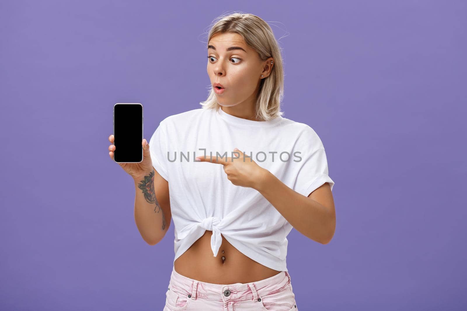 Waist-up shot of impressed and delighted gorgeous european female with blond hair and tattoos looking and pointing at awesome brand new smartphone standing over purple background. Technology concept