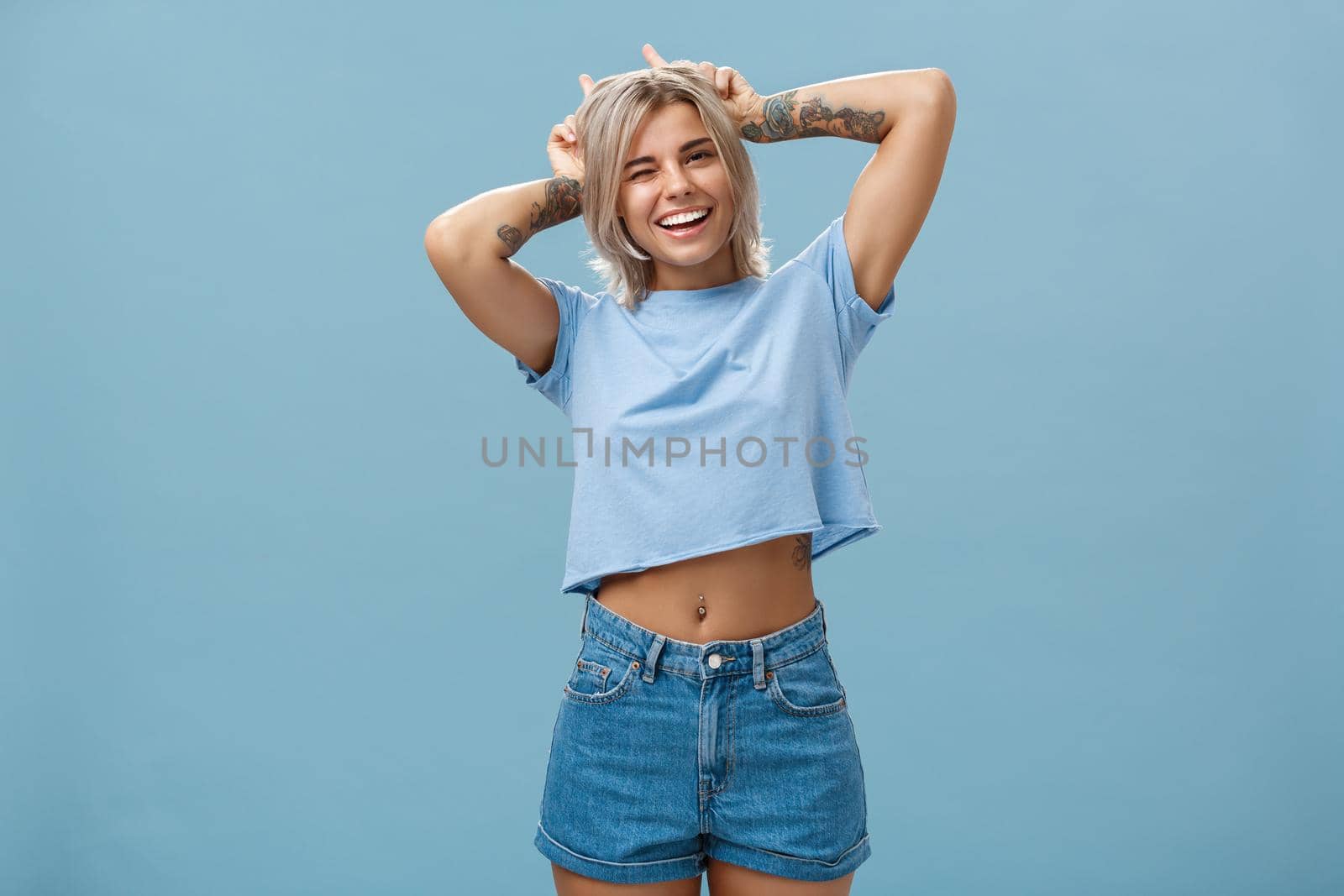 Lifestyle. Studio shot of cute attractive and joyful stylish blond woman with tattoos on arms winking and smiling broadly holding index fingers on head making horns standing in summer outfit over blue wall.