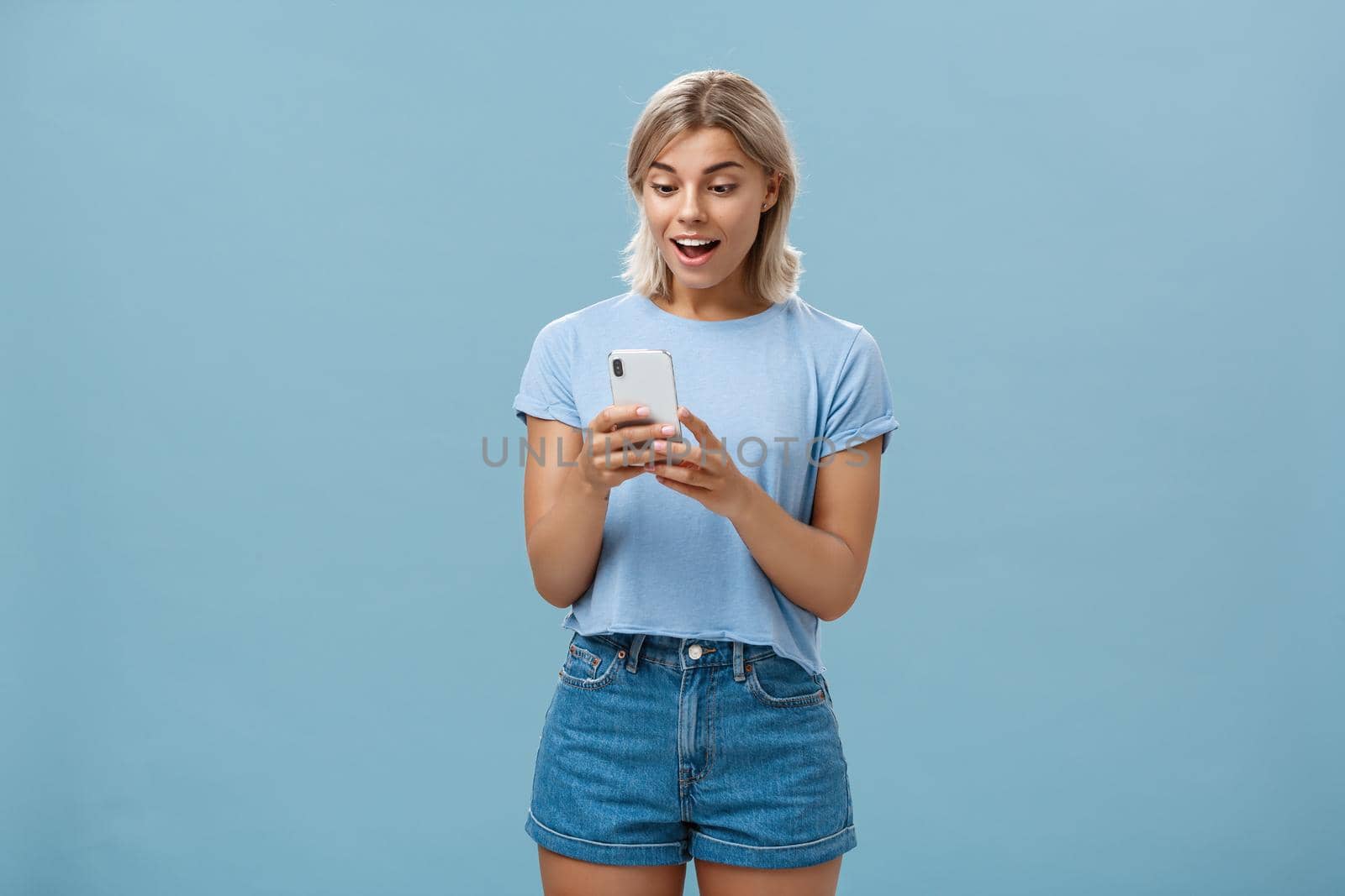 Entertained charming young european female with blond hair in casual outfit holding smartphone being satisfied with great camera and photos gasping and gazing amazed at screen over blue wall. Technology concept