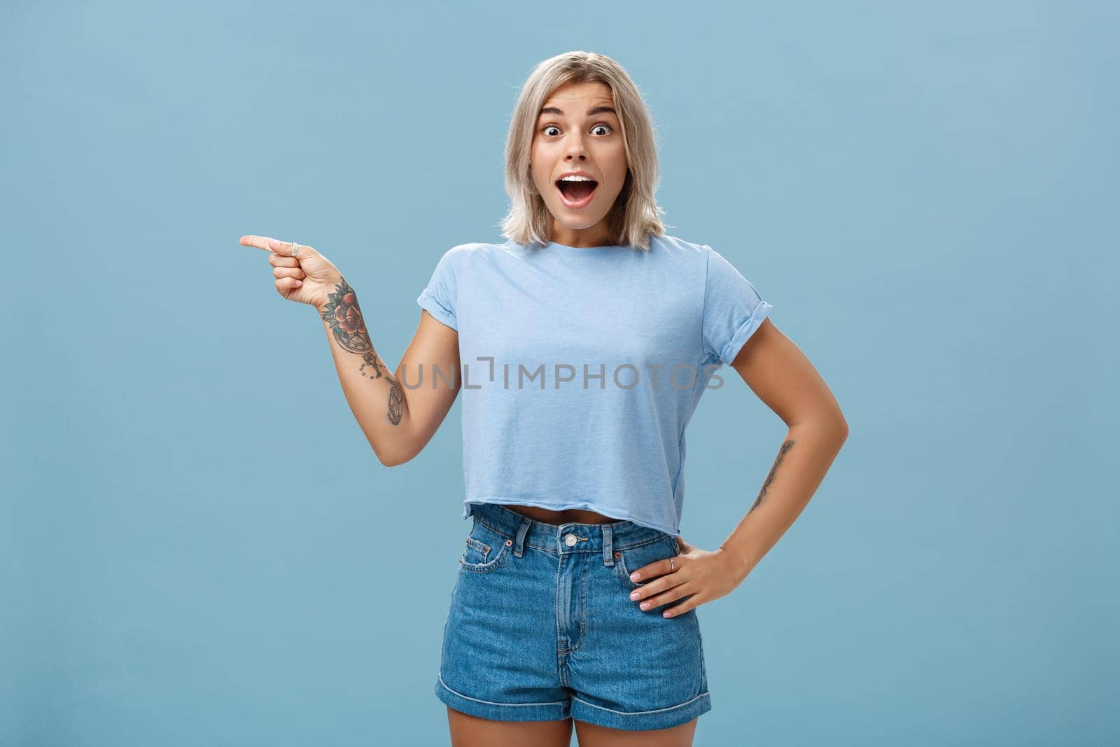 Studio shot of amazed impressed good-looking sportswoman in trendy t-shirt with tattoos opening mouth with enthusiasm from surprise holding hand on waist and pointing left over blue background. Emotions concept