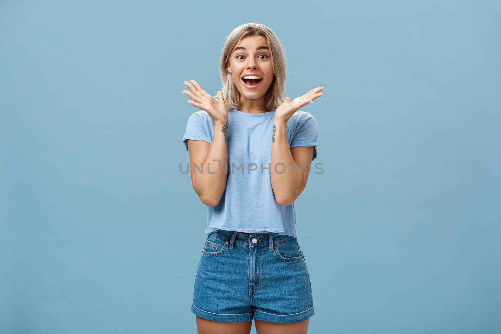 Thrilled enthusiastic happy woman in trendy t-shirt and shorts clapping hands from amazement smiling broadly and gazing with admiration at camera being surprised and joyful over blue wall by Benzoix