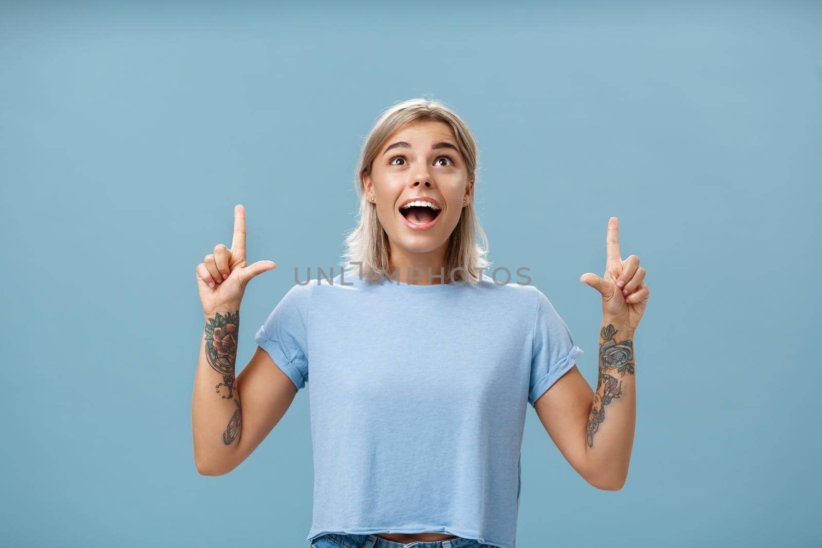 Studio shot of impressed speechless happy good-looking woman with tattoos on arms dropping jaw from amazement and joy gazing fascinated and pointing up standing over blue background. Emotions concept