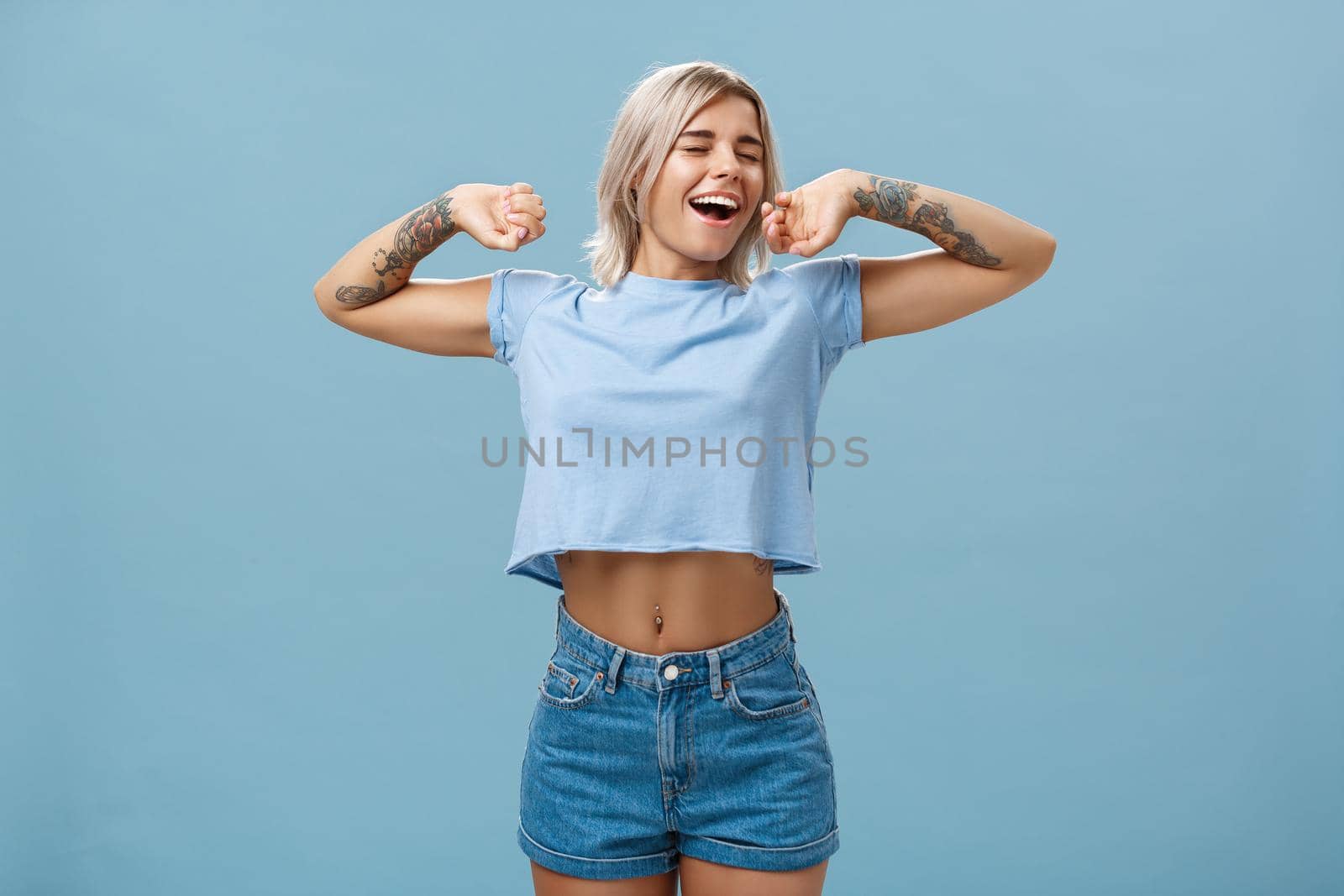 Great feelings after nap. Satisfied carefree and relaxed attractive athletic and stylish female with tattooed arms stretching hands yawning and smiling with closed eyes and pleased look after sleep. Emotions concept