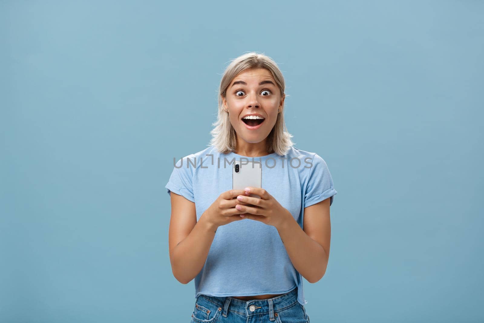 Lifestyle. Indoor shot of amazed attractive blonde female coworker in casual t-shirt gasping from amazement and happiness being surprised staring surprised at camera holding smartphone receiving awesome news.