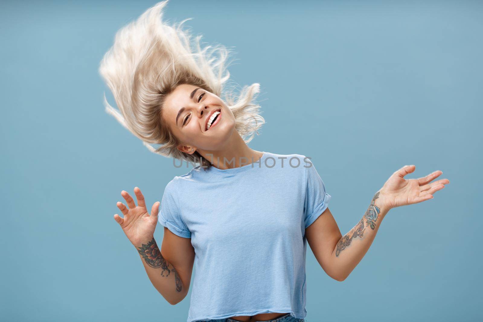 Waist-up shot of artistic and creative attractive carefree blonde female waving head and hair joyfully dancing and jumping having fun smiling broadly over blue background. Emotions and people concept