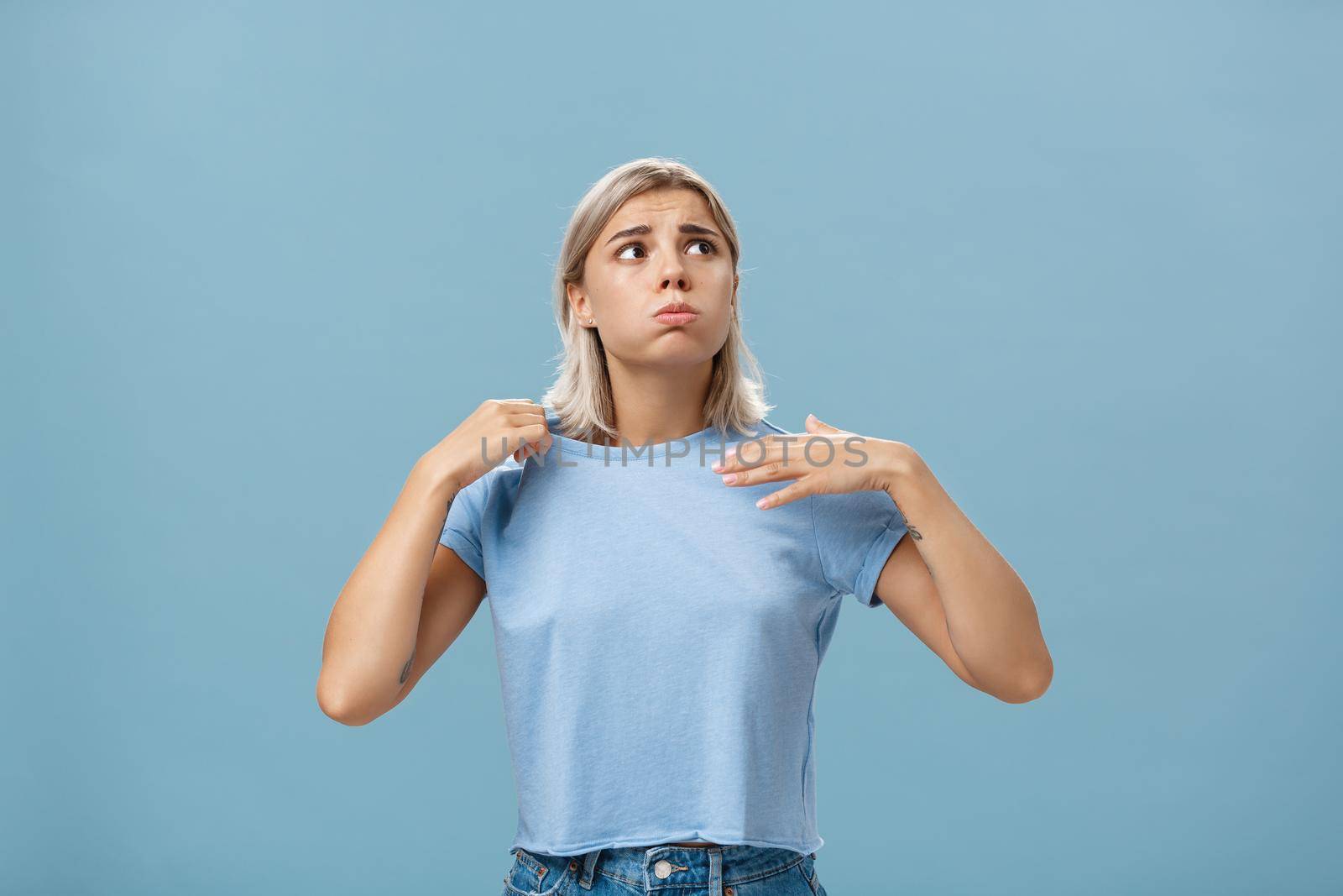 Girl feeling discomfort from heat standing over blue background in fug breathing out and frowning looking up at sun suffering from hot weather waving with t-shirt to cool. Body language concept