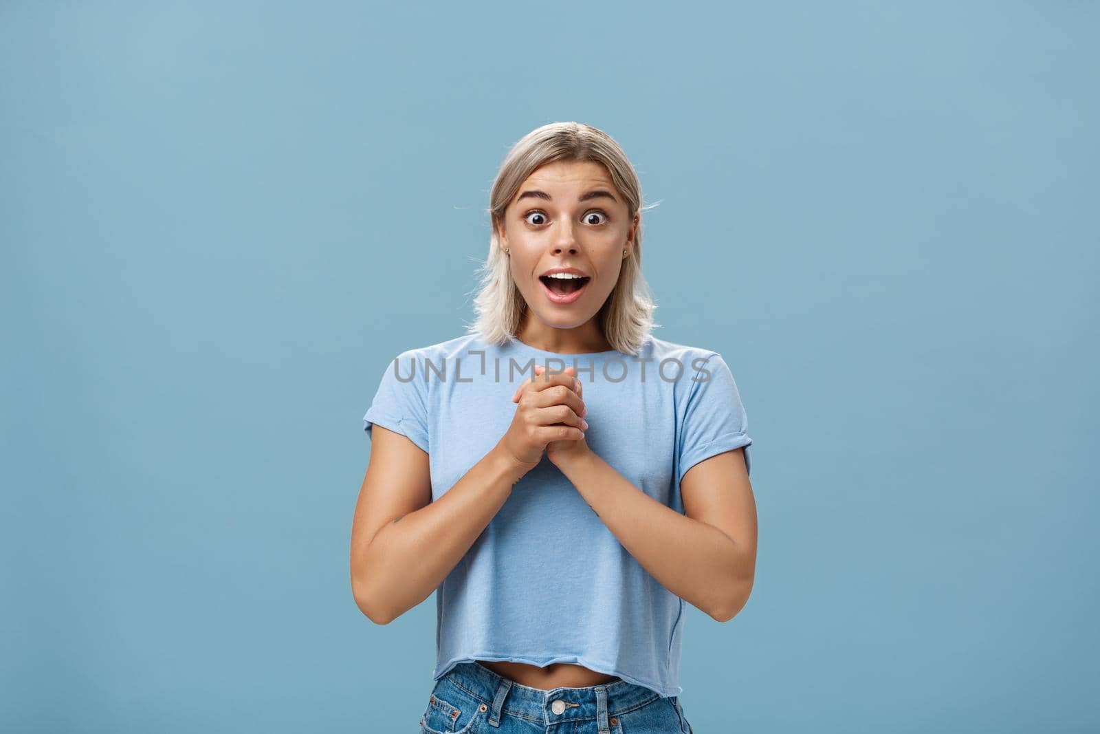 Really is it for me. Portrait of surprised grateful attractive woman with blond haircut clenching hands together on breast opening mouth gasping from admiration and fascination standing over blue wall by Benzoix