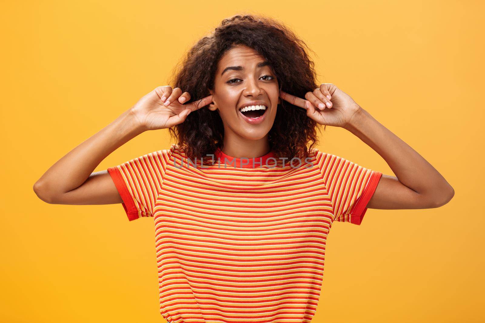 Do not care what saying cannot hear you. Carefree indifferent good-looking african american woman with curly hairstyle closing ears with index finger singing lalala during argument behaving childish.