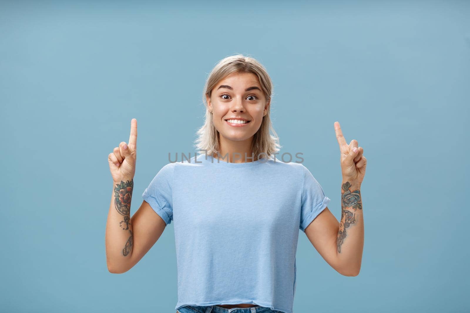 Lifestyle. Waist-up shot of excited and thrilled happy gorgeous blonde with short hairstyle and tattoos on arms smiling joyfully from amazement pointing up with raised index fingers posing over blue background.