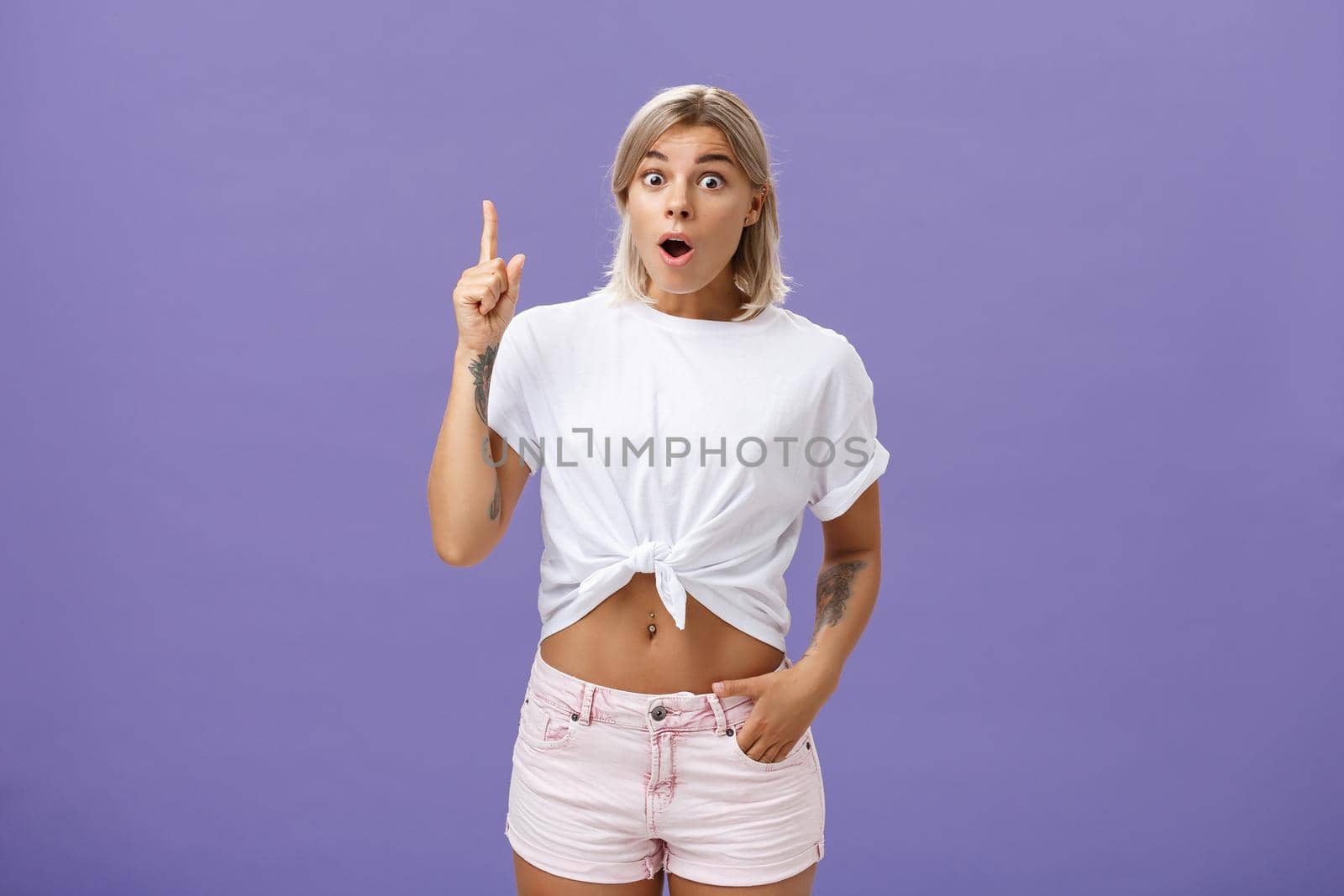 I have got idea. Thrilled and excited attractive stylish feminine woman with tattoos popping eyes folding lips, gasping raising index finger up in eureka gesture having excellent idea over purple wall by Benzoix