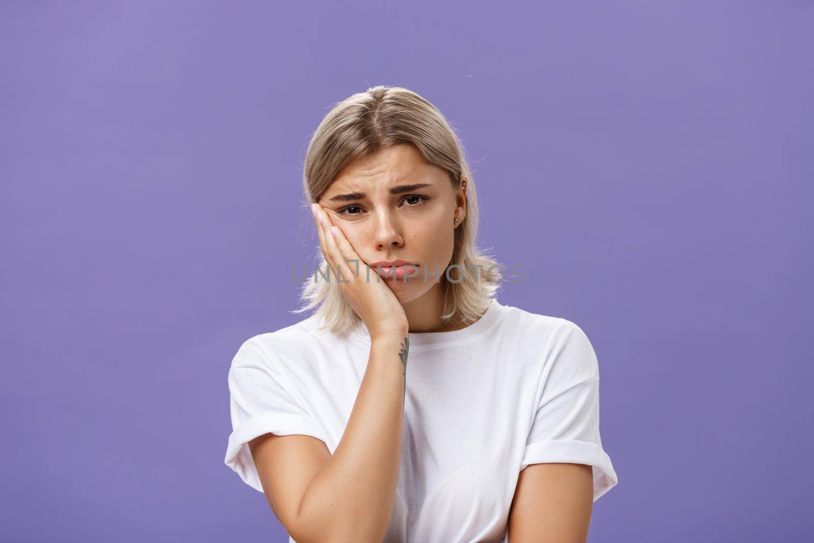 Waist-up shot of unhappy miserable and sad cute blonde female in white casual t-shirt pursing lips leaning face on palm and frowning from disappointement and regret over purple background by Benzoix