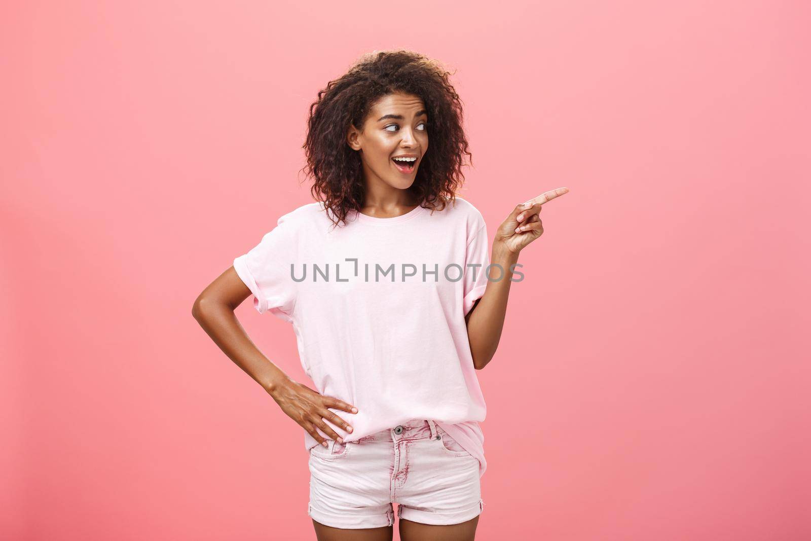 Hey wanna go there. Portrait of intrigued pleased good-looking stylish african woman on vacation wearing summer clothes pointing and gazing right with curious look over pink background by Benzoix