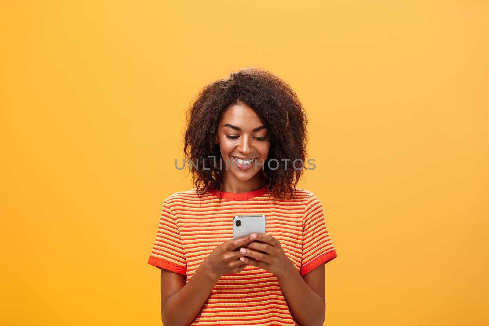 Typing answer to party invitation via smartphone message app holding device over chest smiling joyfully at gadget screen, texting or playing funny game, waste time in subway over orange wall by Benzoix