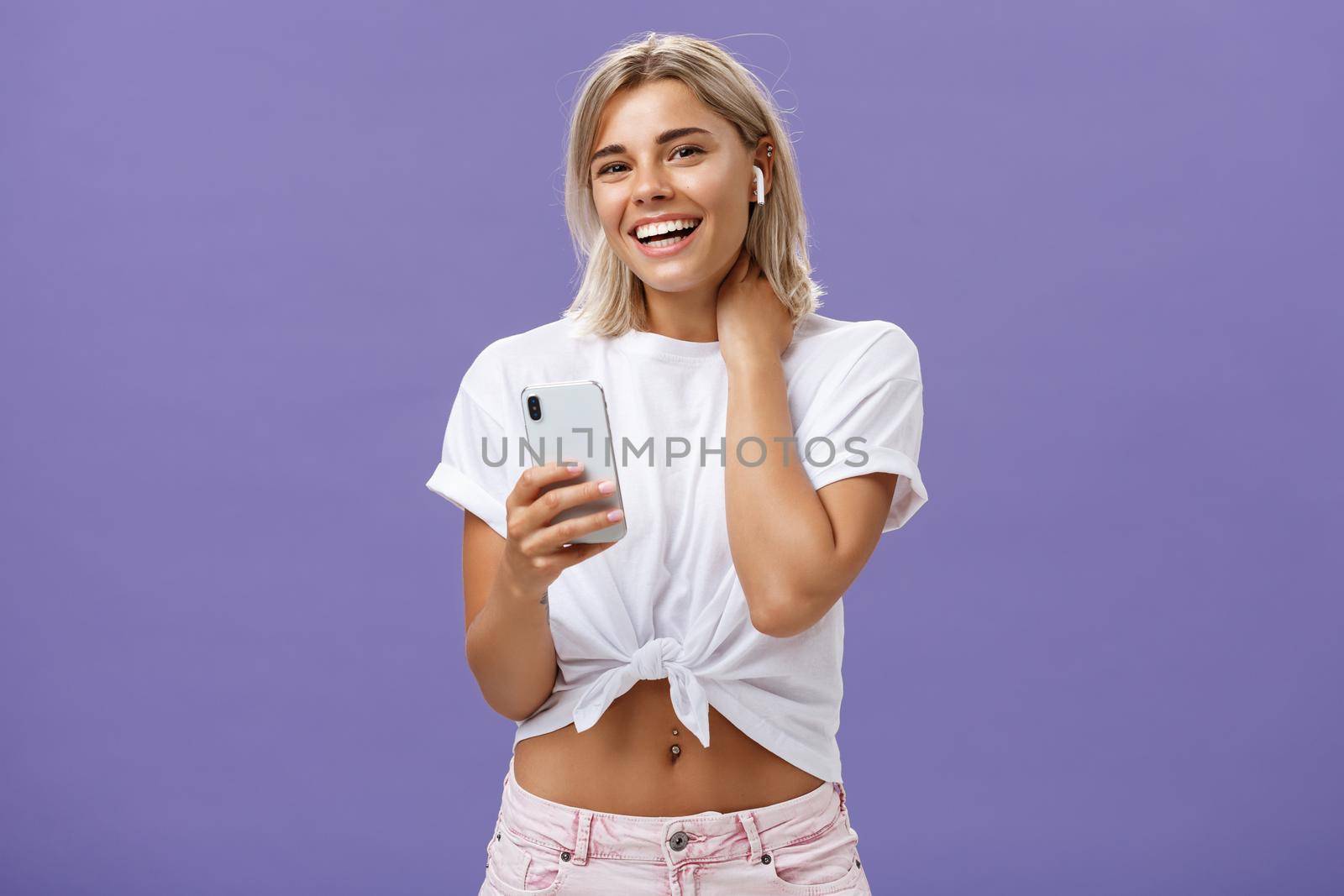 Shy attractive and stylish sociable woman in white t-shirt touching neck timid and awkward smiling broadly wearing wireless earphones while listening songs with smartphone over purple wall. Technology concept