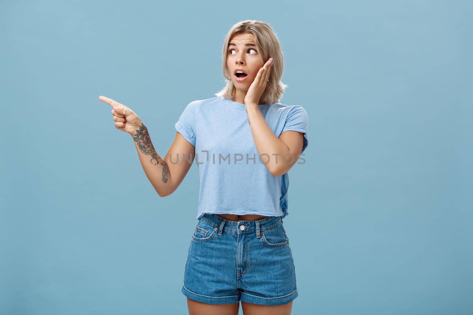 Studio shot of nervous shocked good-looking young woman with fair hair and tatooed arm gasping pointing and looking left with worry holding hand on cheek posing over blue background. Emotions and advertisement concept