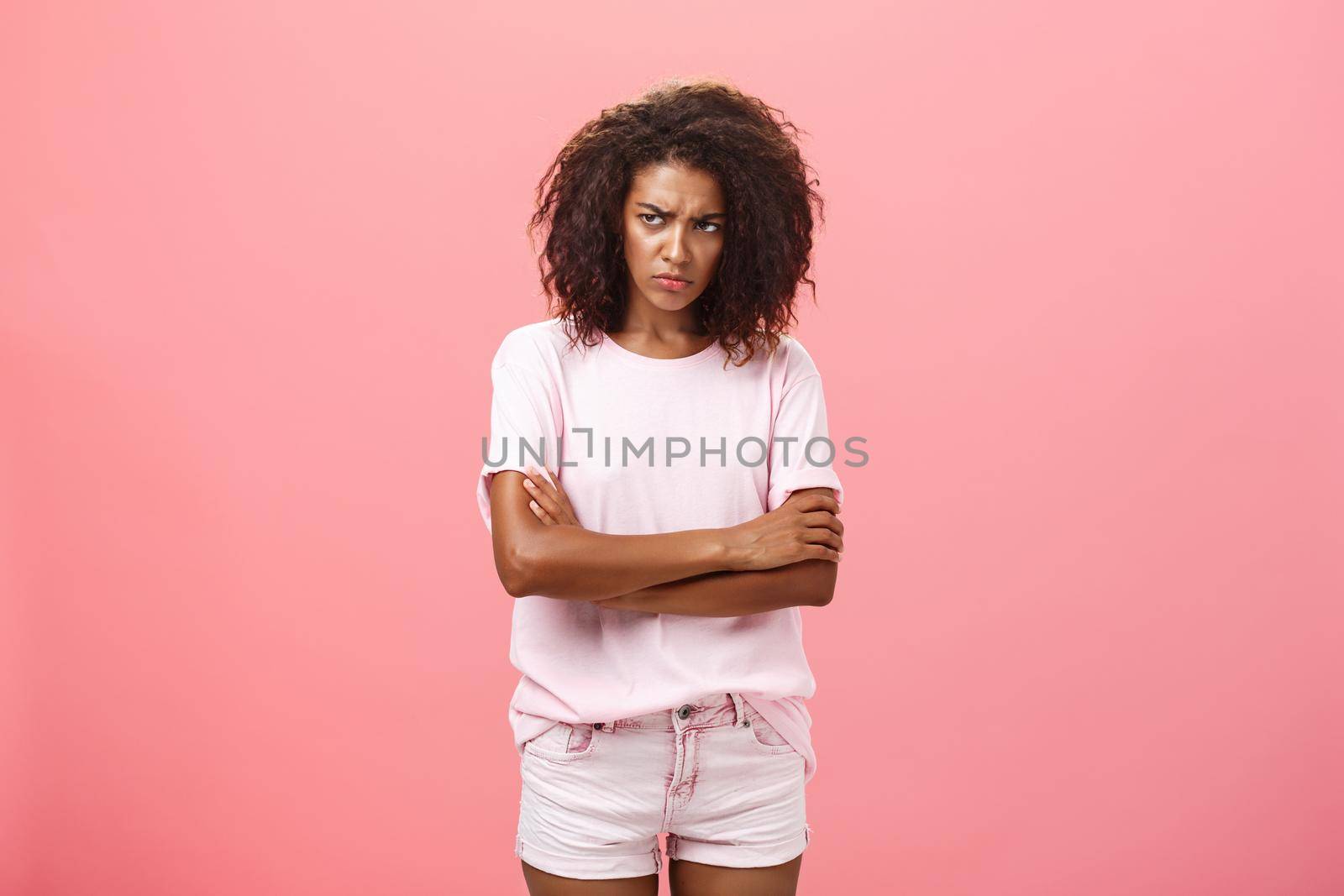 Portrait of offended gloomy sad african american female friend crossing arms on chest in protection gesture frowning looking from under forehead with insult looking envious and angry over pink wall by Benzoix