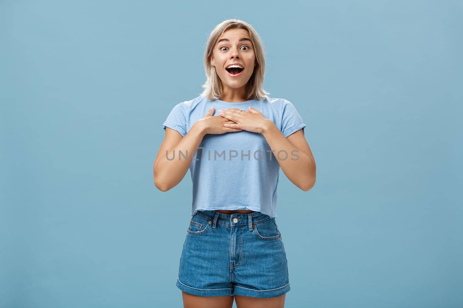 Portrait of amazed and charmed attractive blond female student with tanned skin in denim shorts and summer t-shirt holding palms on breast gasping and smiling joyfully being grateful and pleased by Benzoix