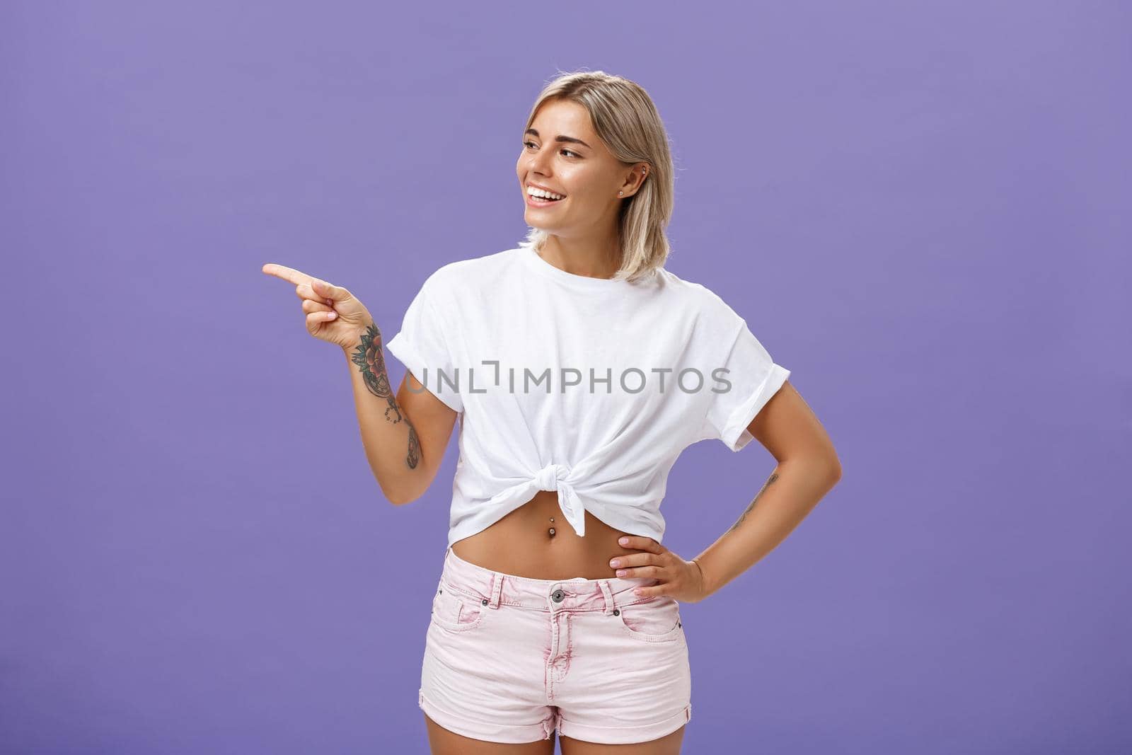 Hey nice job. Pleased carefree good-looking and confident female shop manager in stylish outfit holding hand on waist pointing and looking left with satisfied and delighted look over purple wall by Benzoix