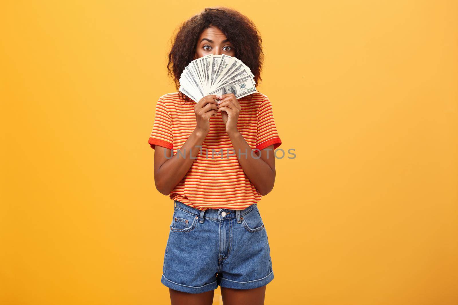 Woman gonna waste lots of cash today. Pleased and delighted rich african american girl with curly hairstyle hiding face behind bunch of money peeking mysteriously at camera winning millions by Benzoix