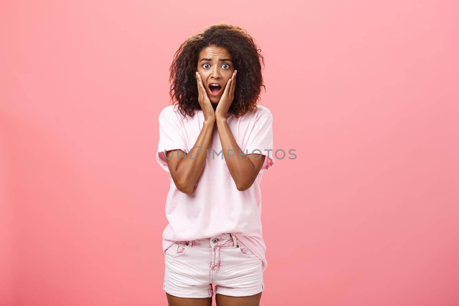 Worried, shocked african american female friend with curly hairstyle in trendy outfit being on vacation seeing scary animal shouting and gasping with opened mouth holding hands on face over pink wall.