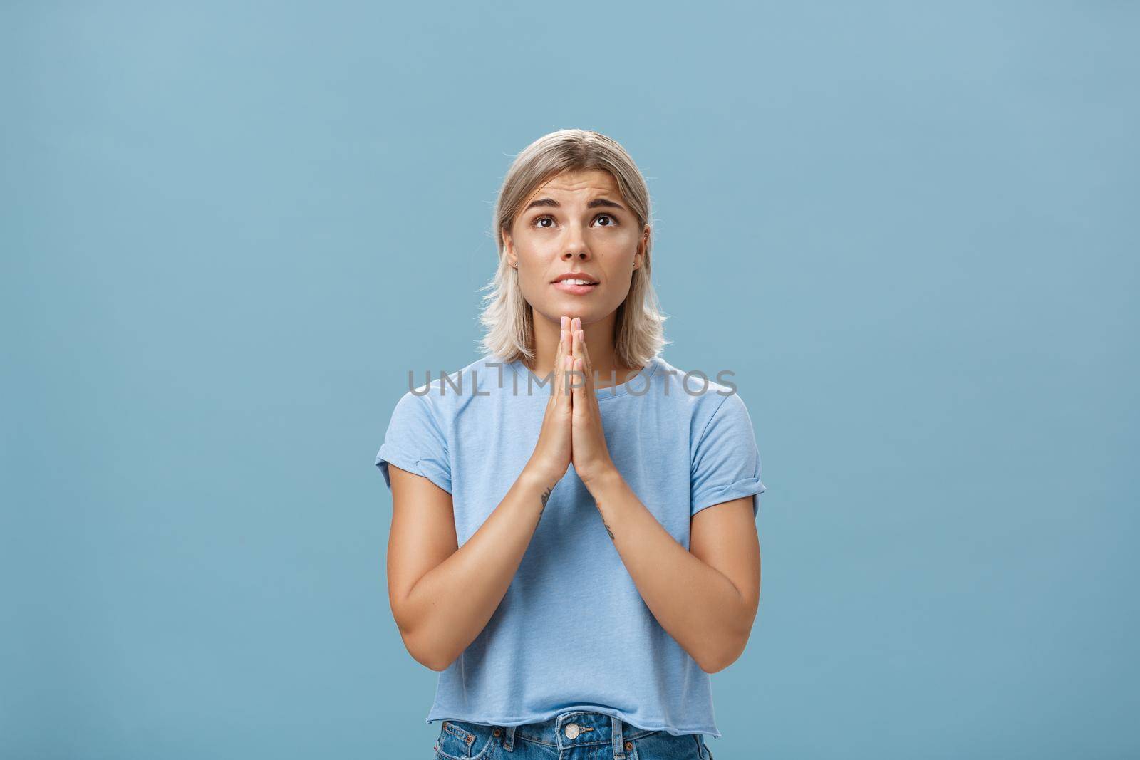 Studio shot of hopeful focused dreamy girl with attractive face and blonde hair holding hands in pray near body looking up hopefully with faith praying or making wish over blue background. Body language concept