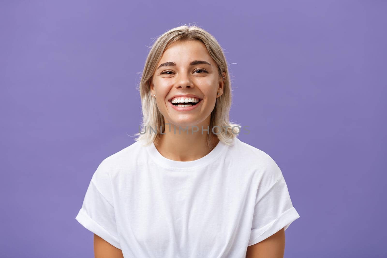 Close-up shot of joyful charming blonde female with delighted and pleased smile standing in white t-shirt over purple background spending time in awesome amusing company. Emotions concept