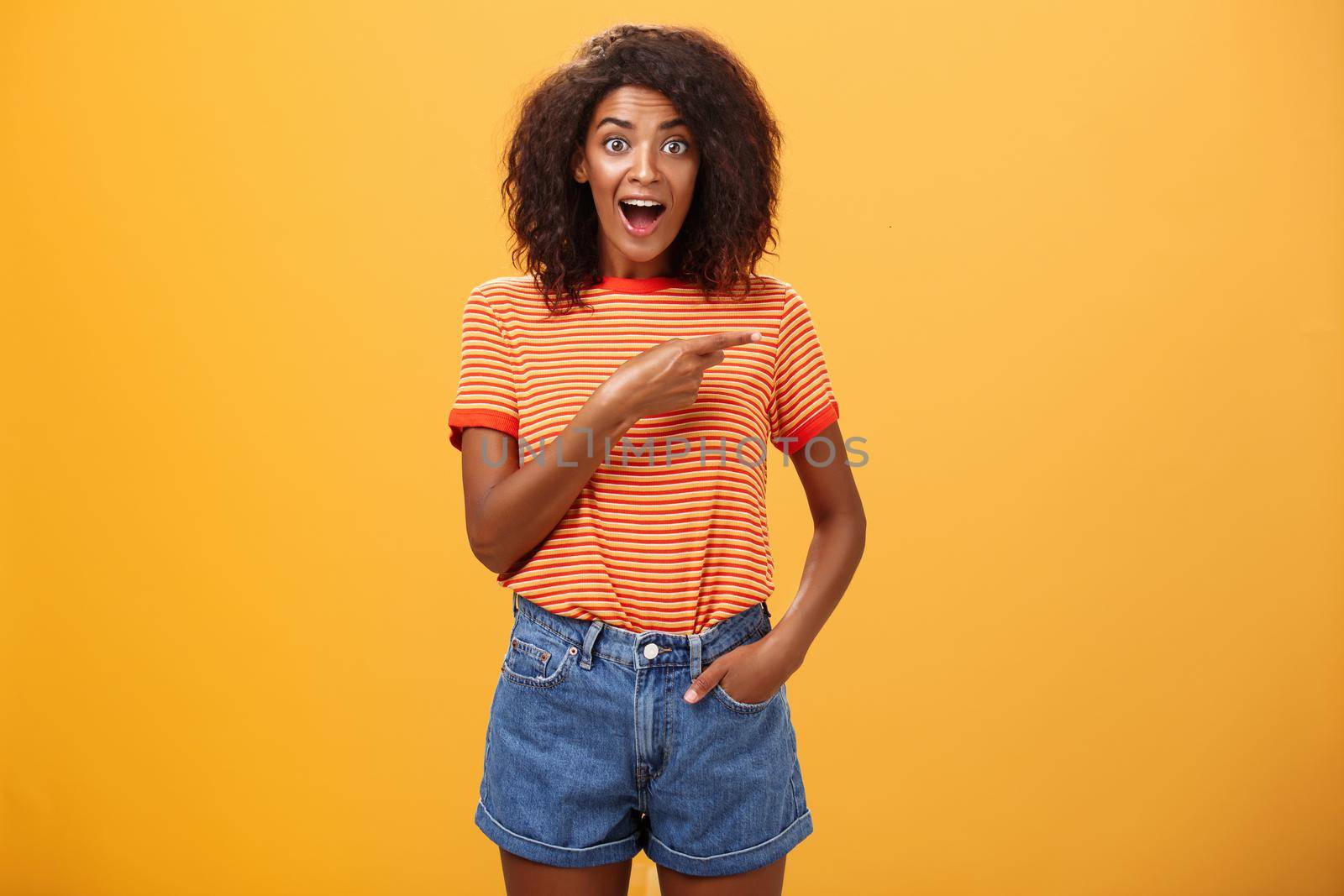 Hey awesme copy space there. Portrait of impressed and surprised enthusiastic young african american female with afro hairstyle pointing right astonished looking energized and curious over orange wall.