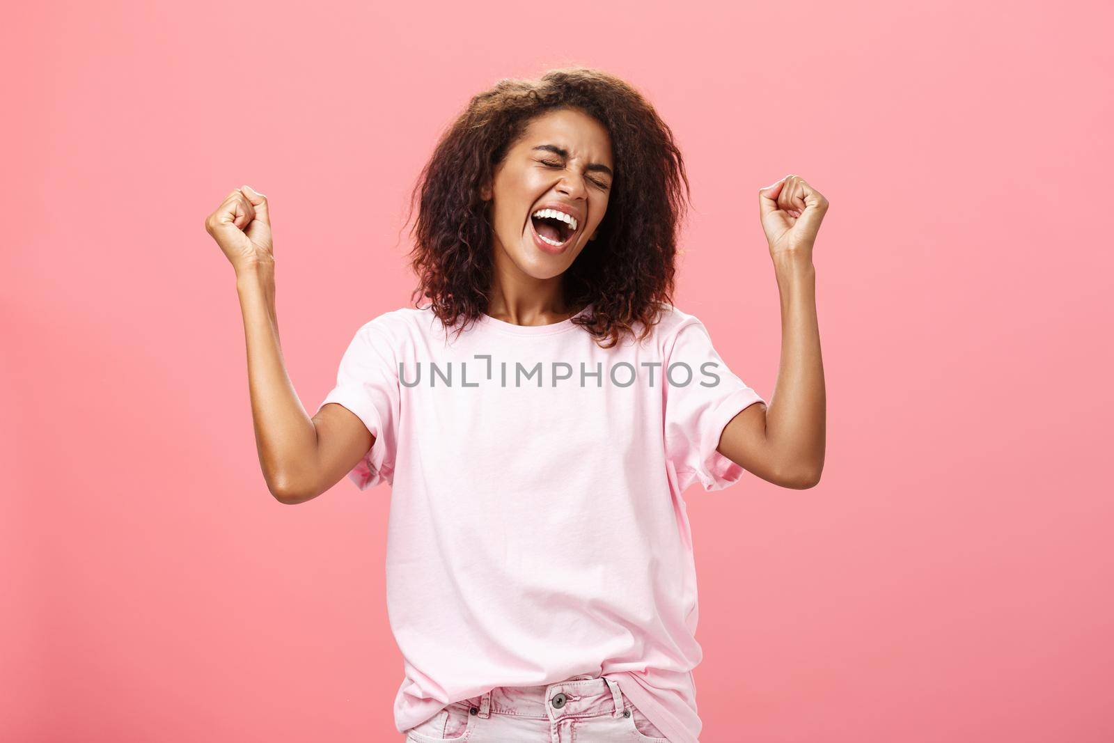 Amazed and happy triumphing african american sportswoman celebrating victory yelling from awesome exciting feelings closing eyes raising fists hight in win gesture standing over pink background. Lifestyle.